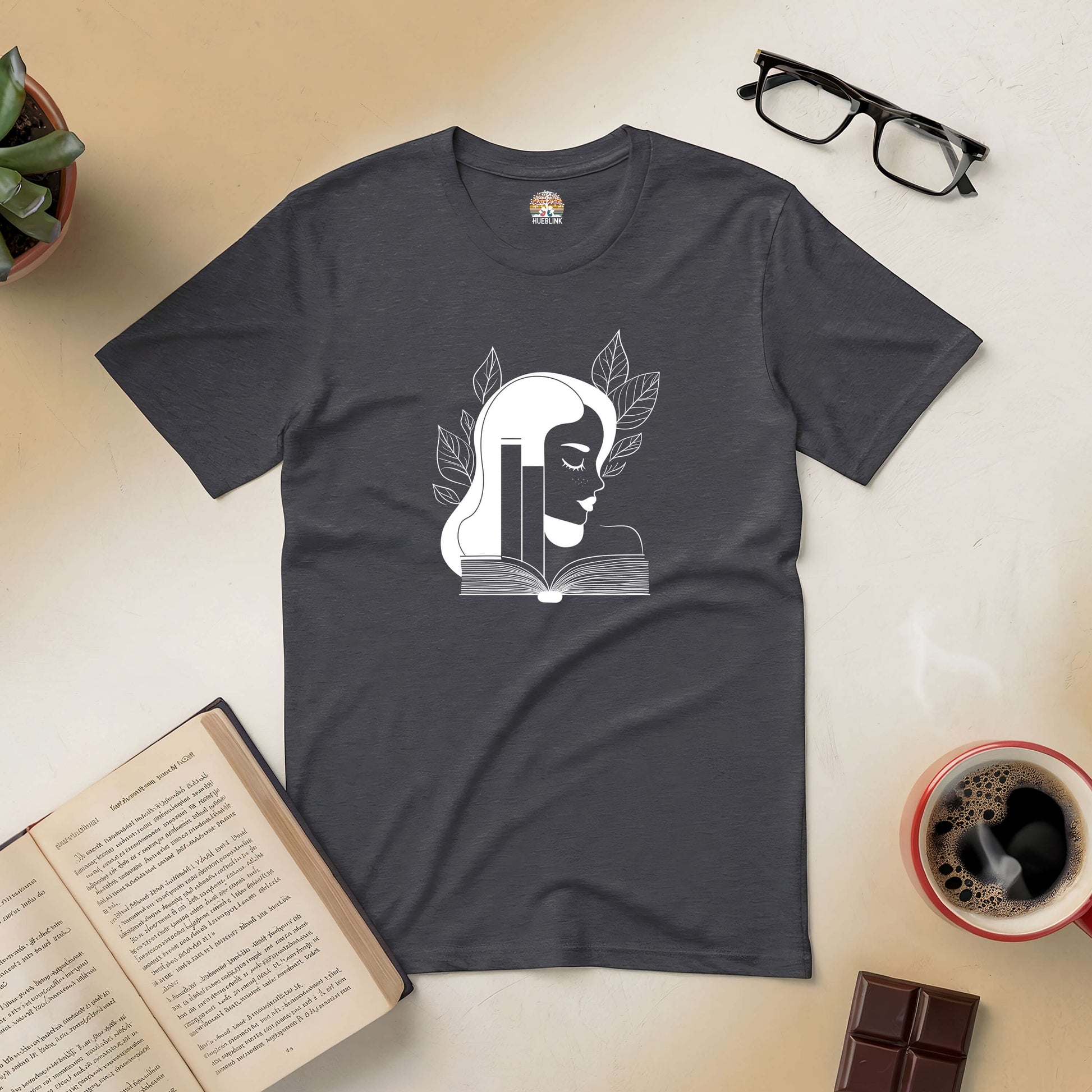 Book-themed tee with elegant design, perfect for book lovers. Gray shirt on table with open book, glasses, coffee, and plant.