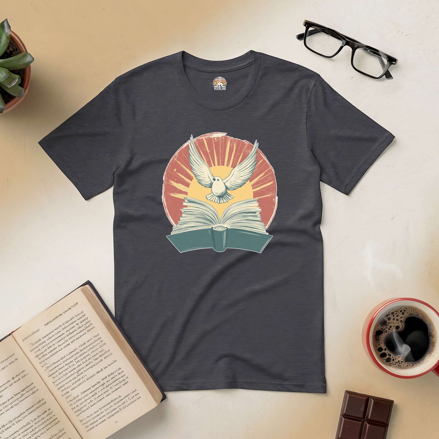 "Boundless Horizons Tee featuring a dove and open book design, surrounded by coffee, glasses, and a book."