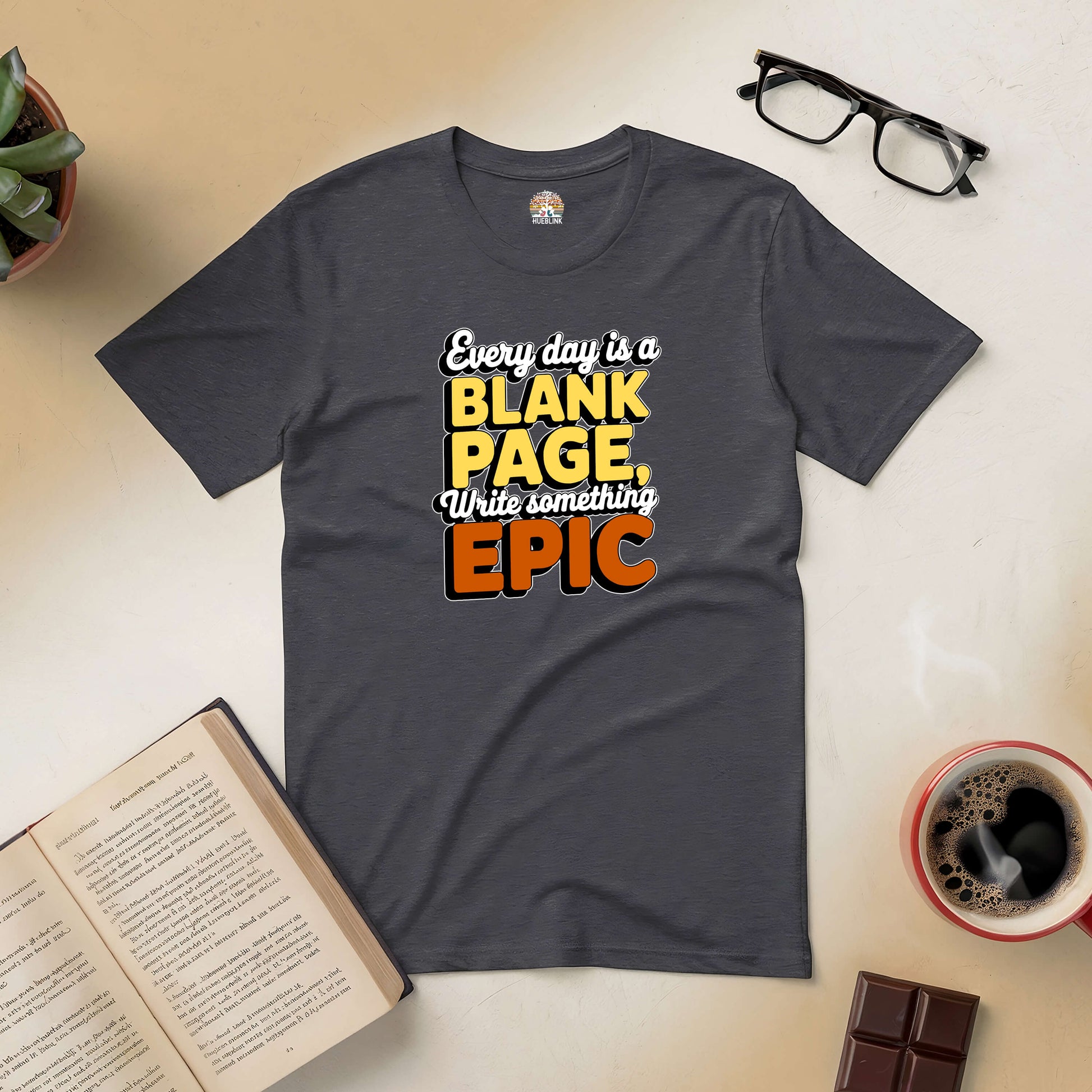 Inspirational tee with "Every Day is a Blank Page, Write Something Epic" design, surrounded by coffee and books.