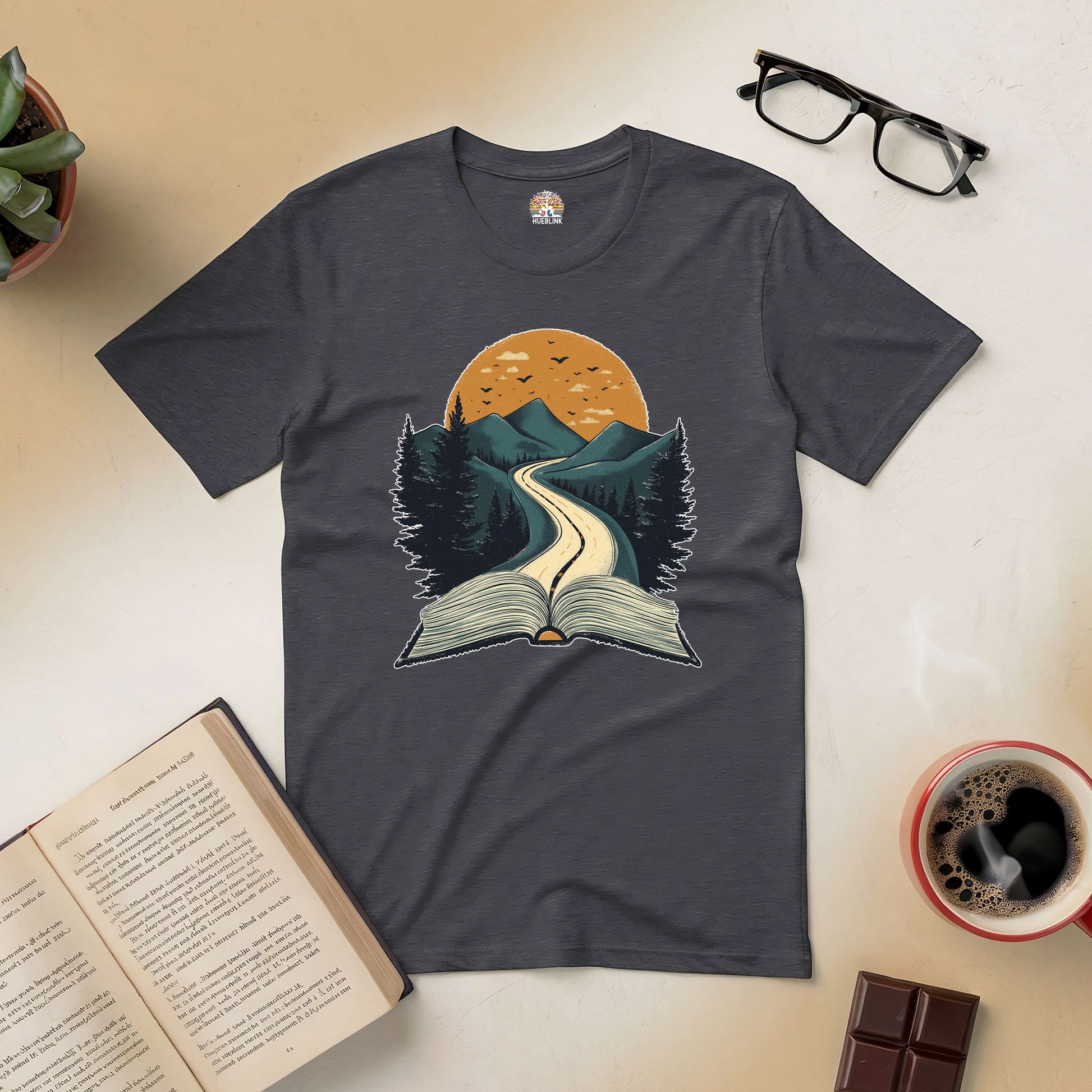 Dark grey Starting Point Tee with open book and mountain path design, surrounded by coffee, glasses, and plants, ideal for readers.