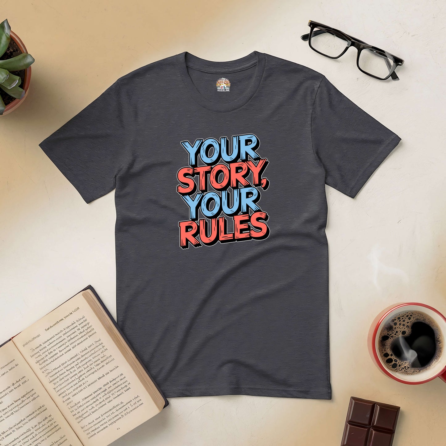 "Your Story, Your Rules Tee promoting individuality and confidence, flat lay with book, glasses, coffee, plant, and chocolate"