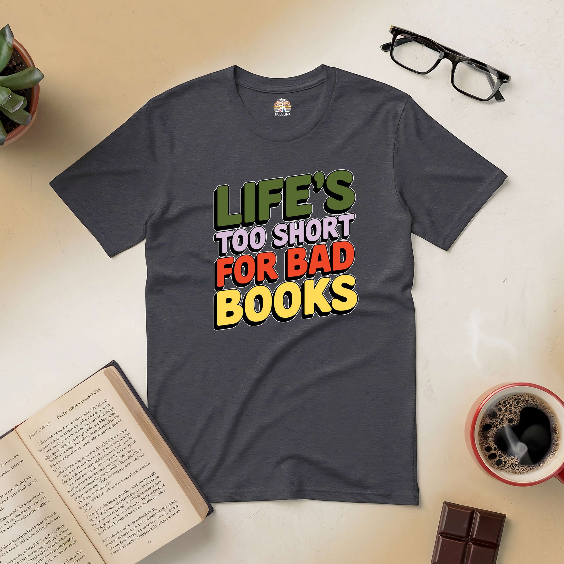 Charcoal tee with "Life's Too Short for Bad Books" design, surrounded by book, glasses, coffee, and chocolate on a table.