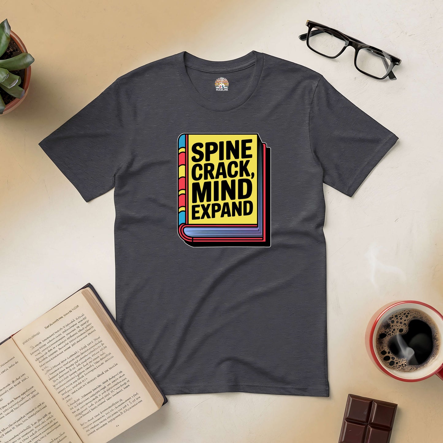 "Spine Crack, Mind Expand Tee with book graphic, celebrating worn pages and open minds, surrounded by glasses, coffee, and an open book."