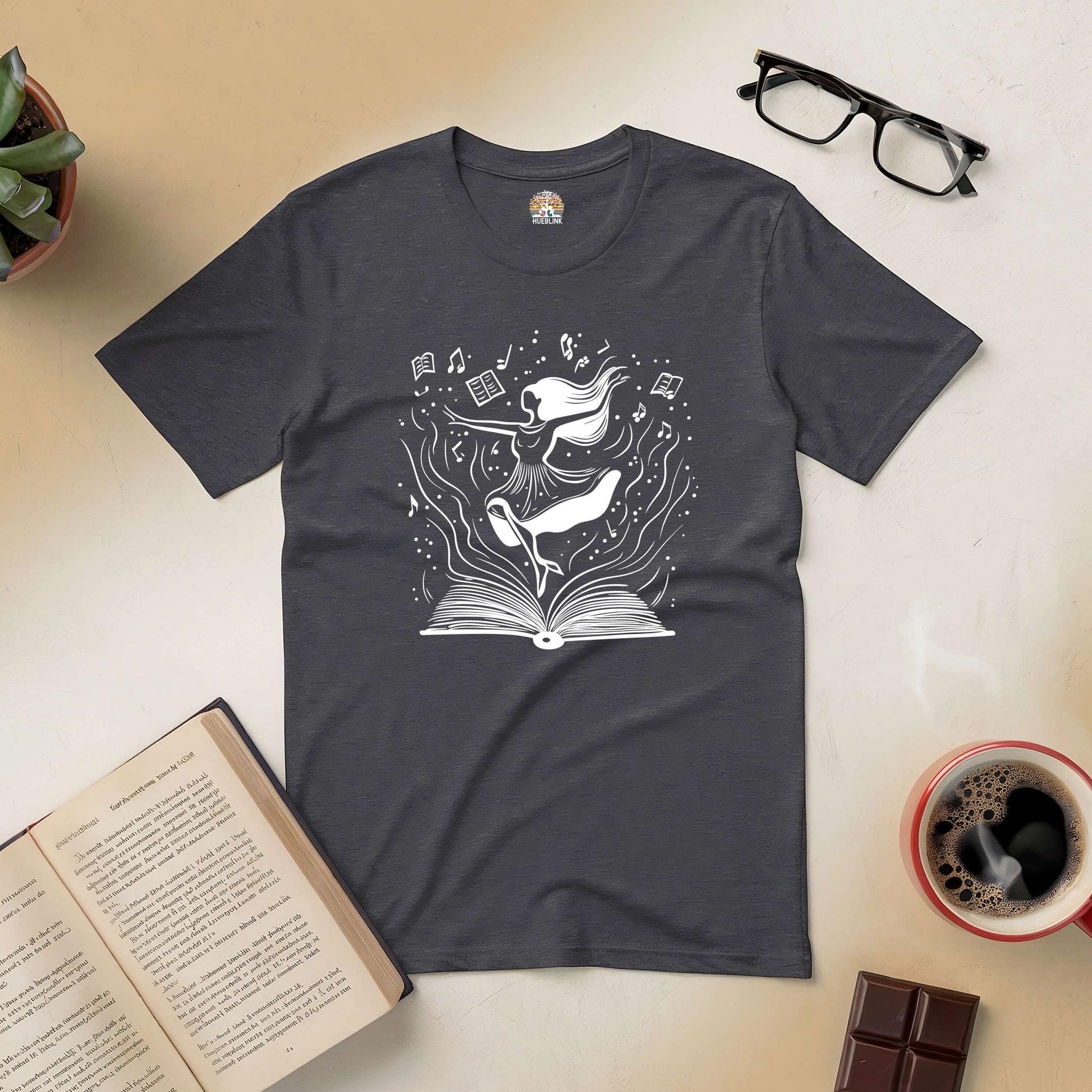 "Dance Through the Pages tee with open book and dancing figures, surrounded by glasses, coffee, chocolate, and a plant."