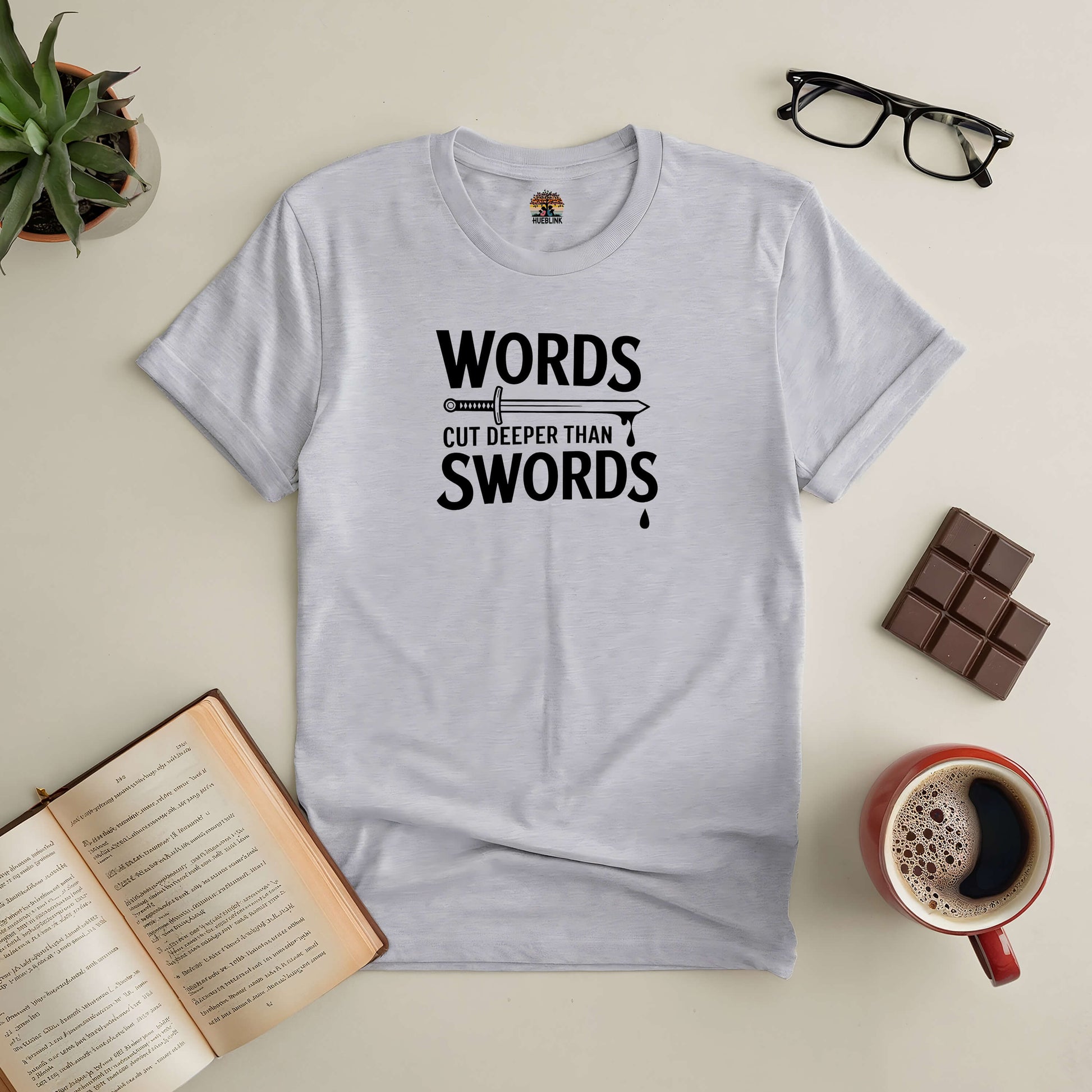 Gray tee with "Words Cut Deeper Than Swords" slogan, surrounded by book, coffee, chocolate, glasses, and plant on a table.