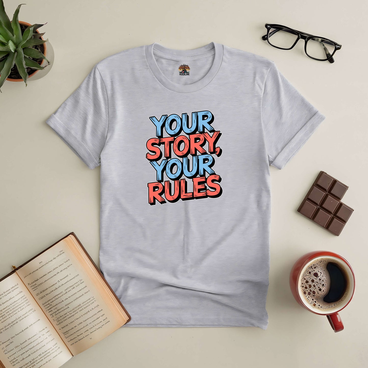 "Your Story, Your Rules Tee promoting individuality and self-expression lies flat with accessories like glasses and coffee"