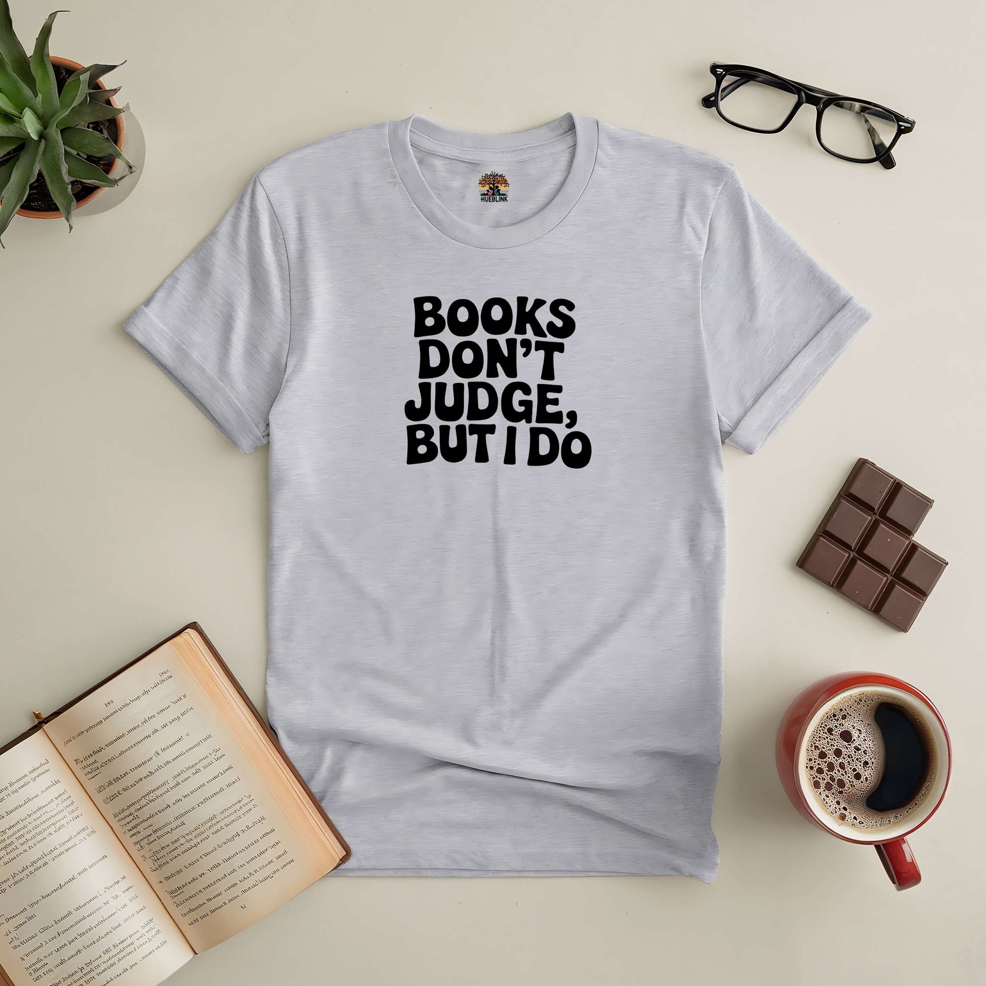 Gray "Books Don't Judge, But I Do" tee with open book, coffee, chocolate, and glasses on a table.