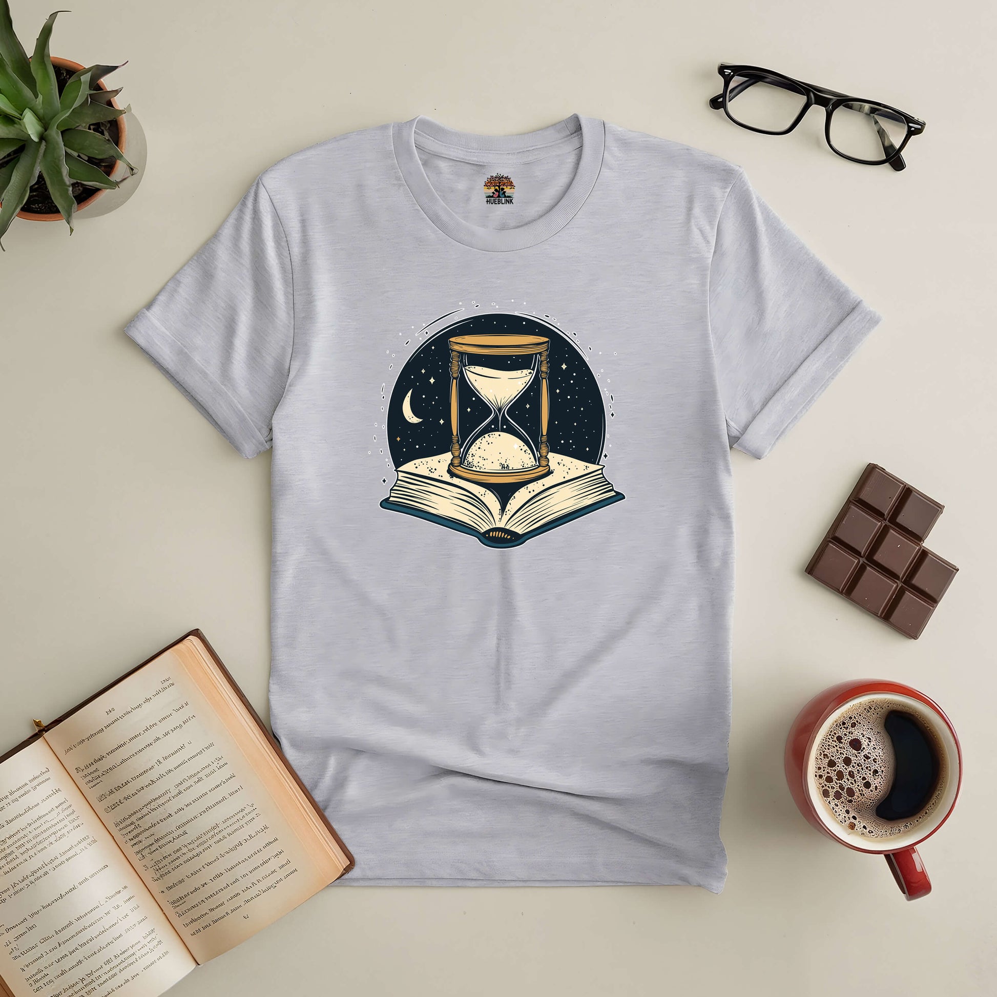 "Chronicles of Time and Space Tee with hourglass and open book design, surrounded by coffee, chocolate, and greenery"