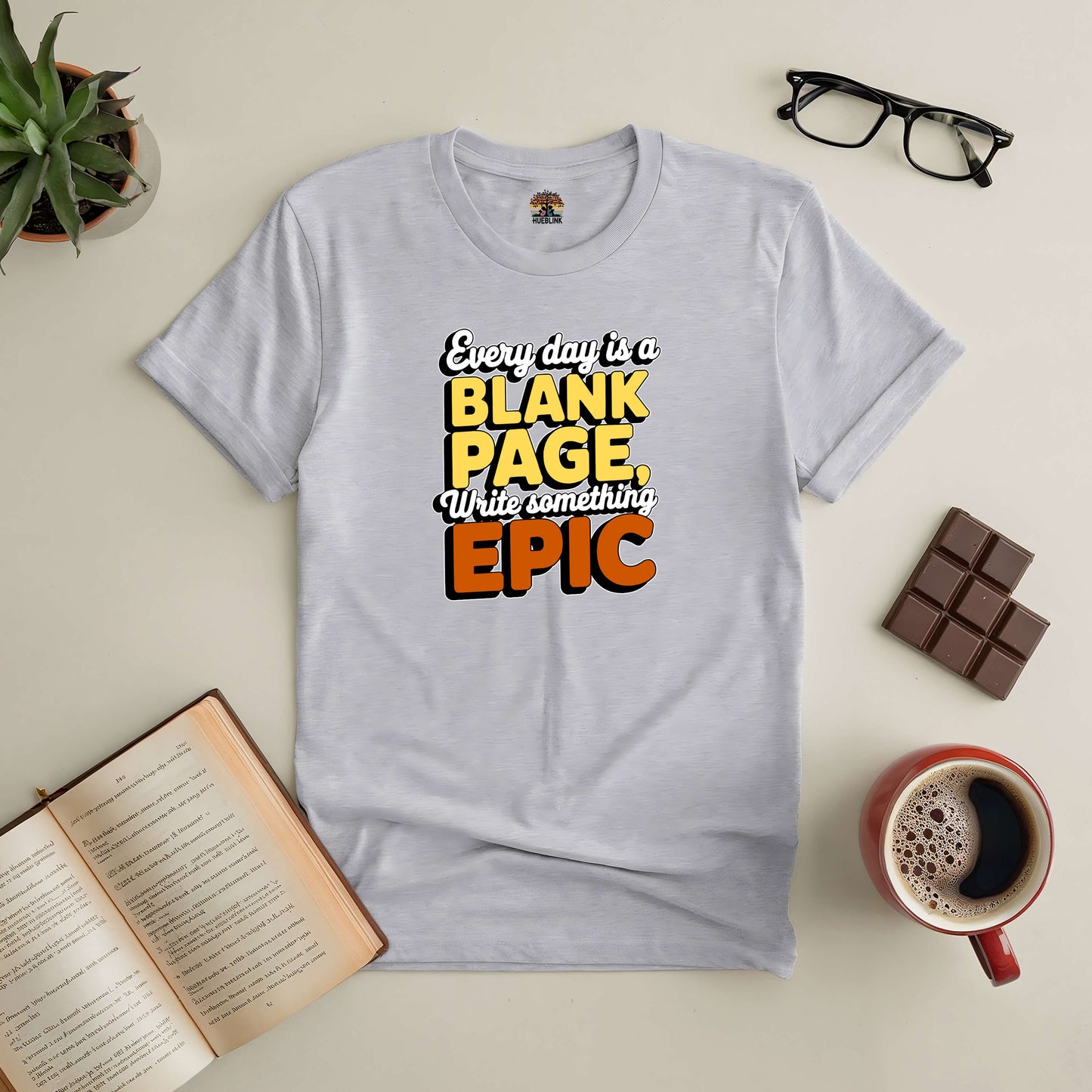 Inspirational gray t-shirt with "Every Day is a Blank Page, Write Something Epic" text, styled with book, coffee, and glasses.