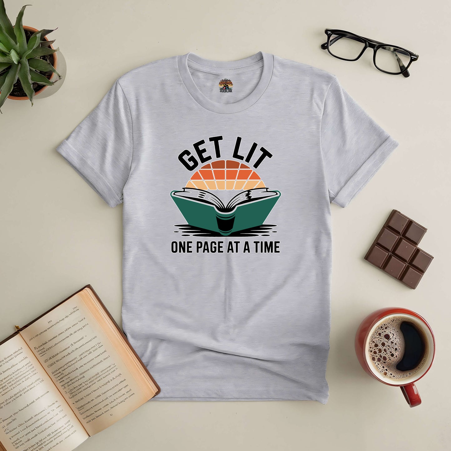 White "Get Lit: One Page At A Time" tee for book lovers, surrounded by coffee, chocolate, open book, glasses, and a plant.