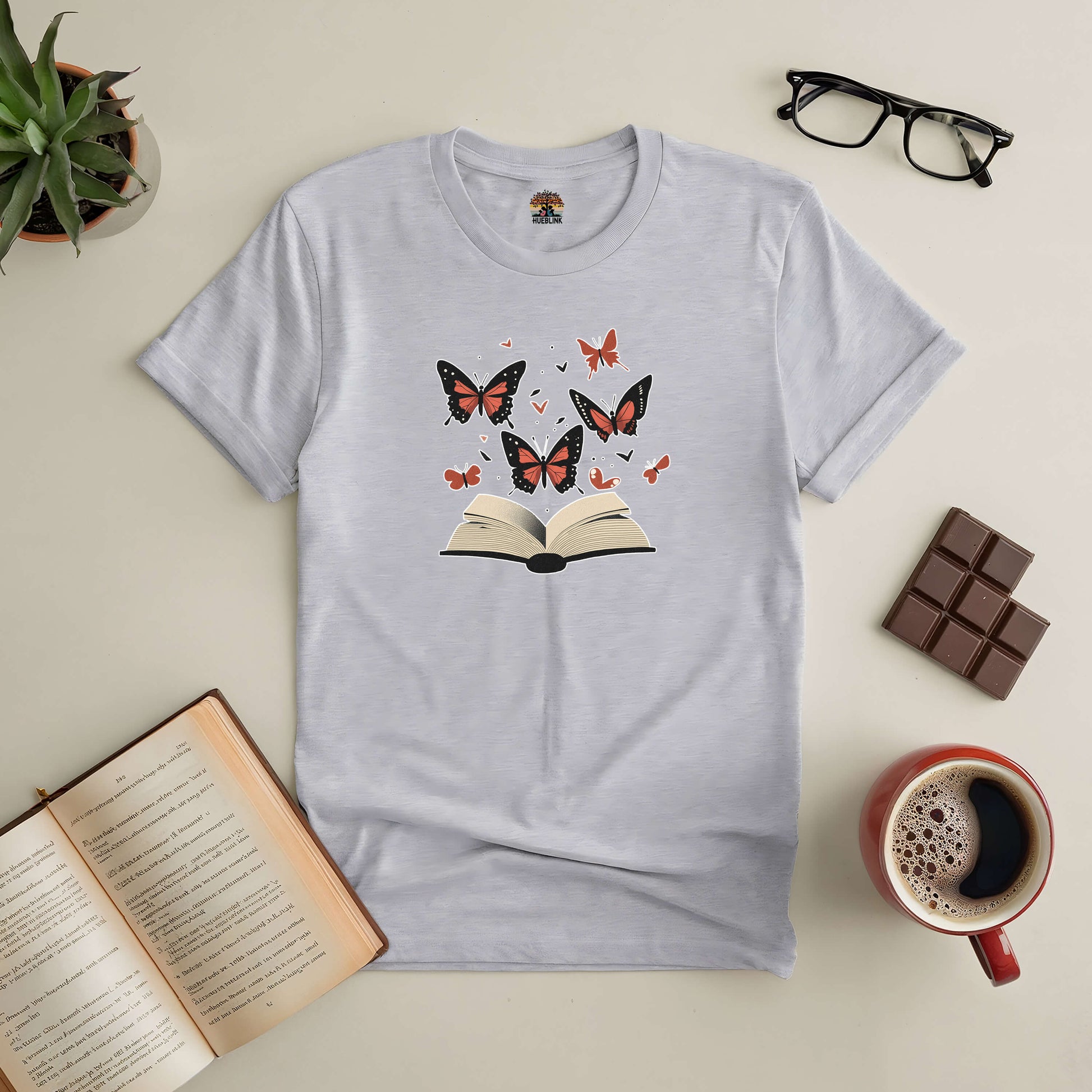 "Books: Where Stories Take Flight tee with open book and butterflies design, surrounded by coffee, chocolate, and reading glasses"