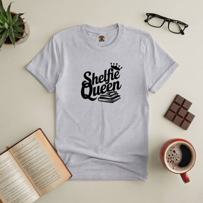 "Shelfie Queen tee with book and crown design, surrounded by an open book, coffee, chocolate, glasses, and a plant on a table."