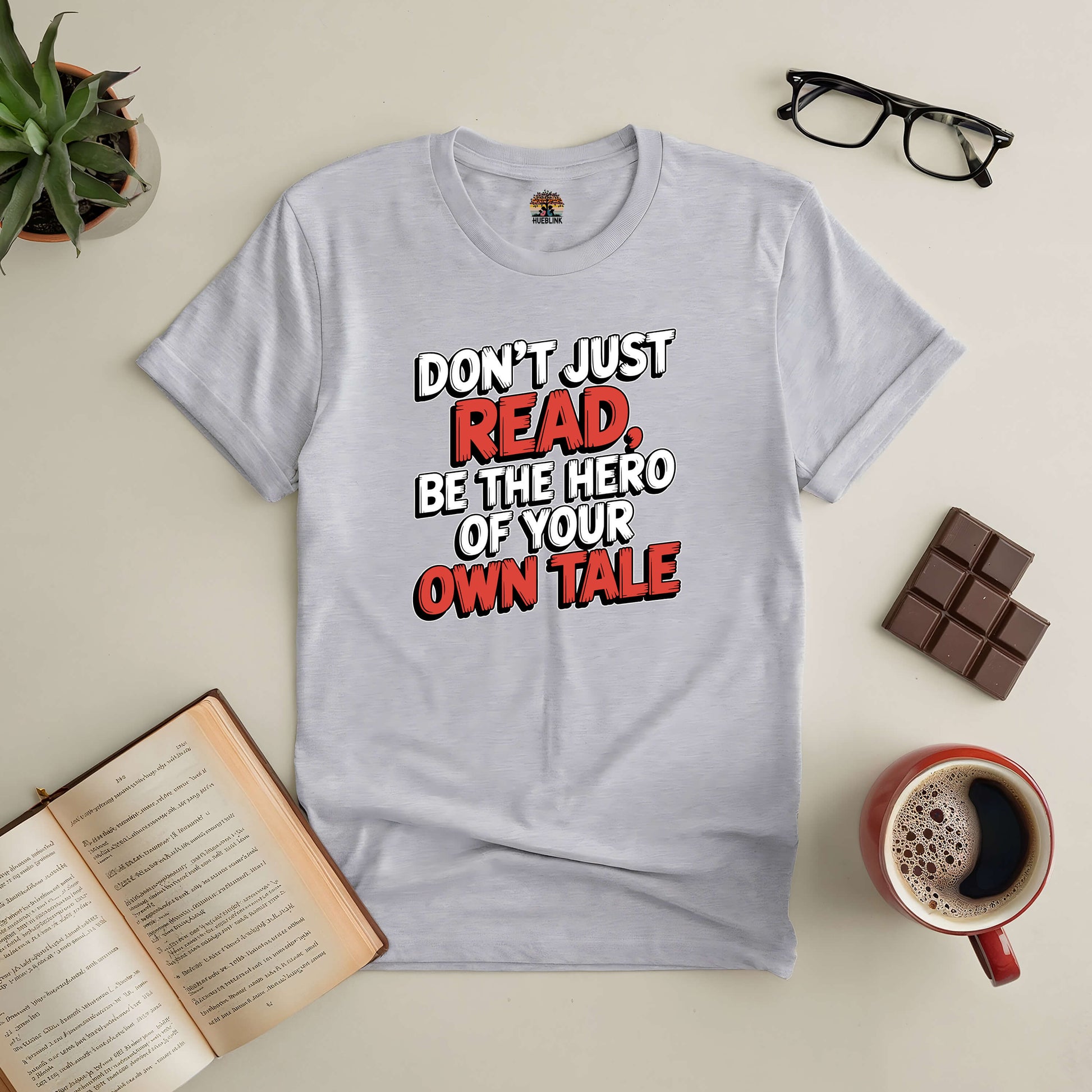 Gray t-shirt with "Don't Just Read, Be the Hero of Your Own Tale" text, surrounded by an open book, glasses, coffee, and chocolate.