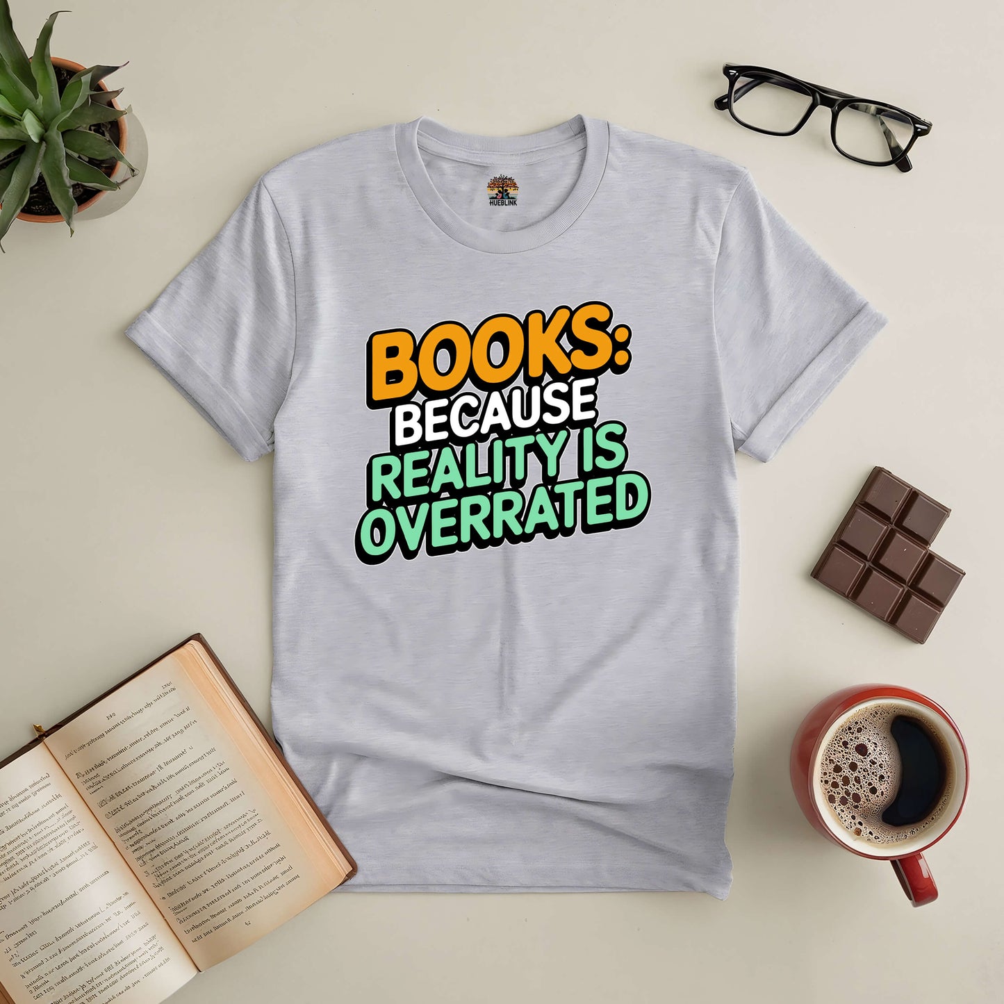 Grey tee with "Books: Because Reality Is Overrated" text, surrounded by an open book, coffee cup, glasses, and a plant.