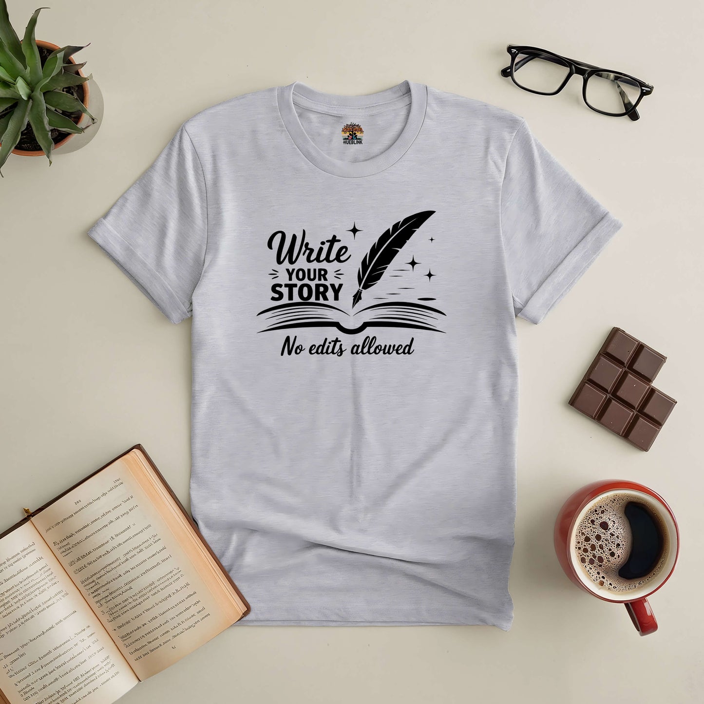 "Write Your Story, No Edits Allowed" tee on a table with coffee, glasses, chocolate, plant, and open book.