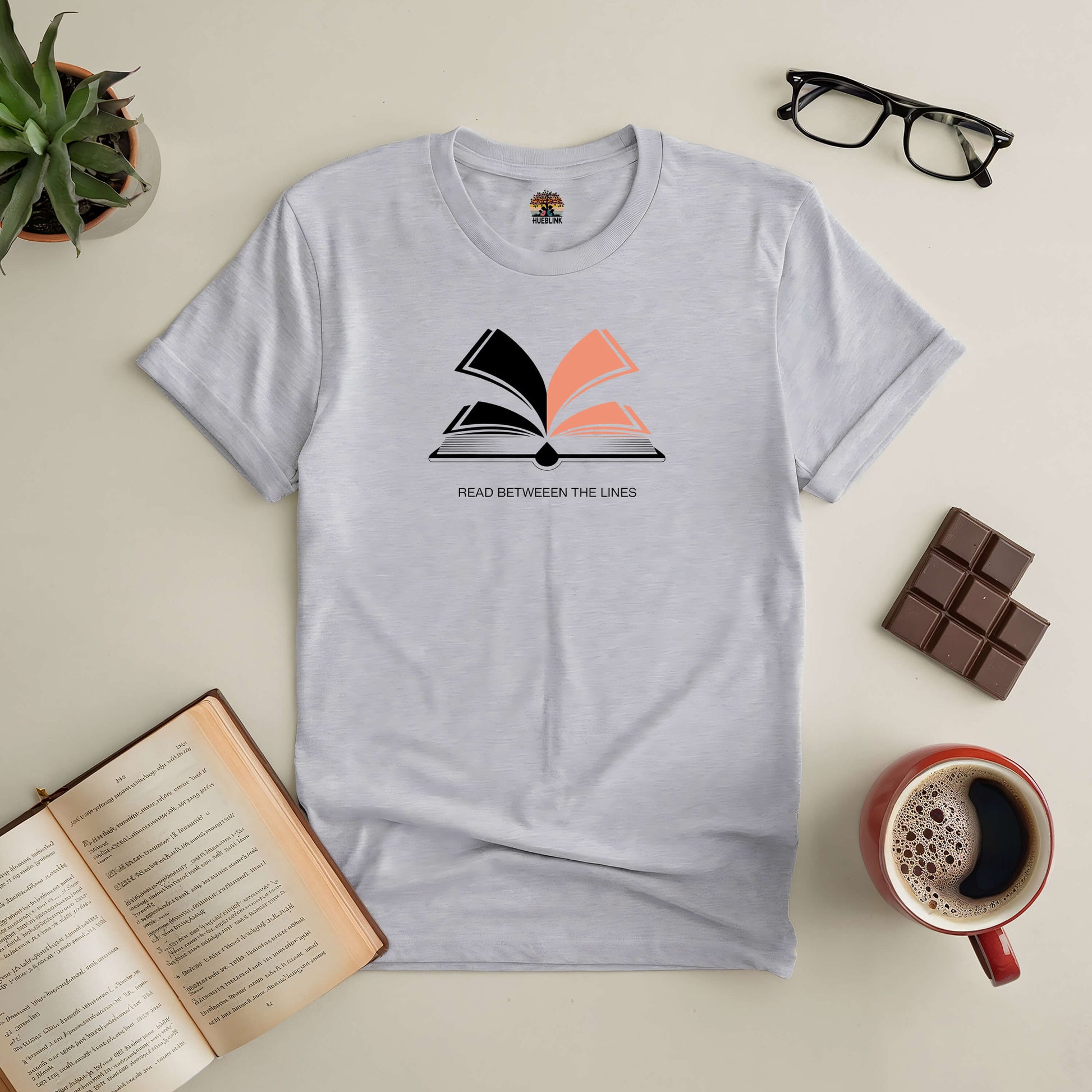 Gray "Read Between The Lines" tee with book graphic, surrounded by coffee, glasses, chocolate, a plant, and an open book.