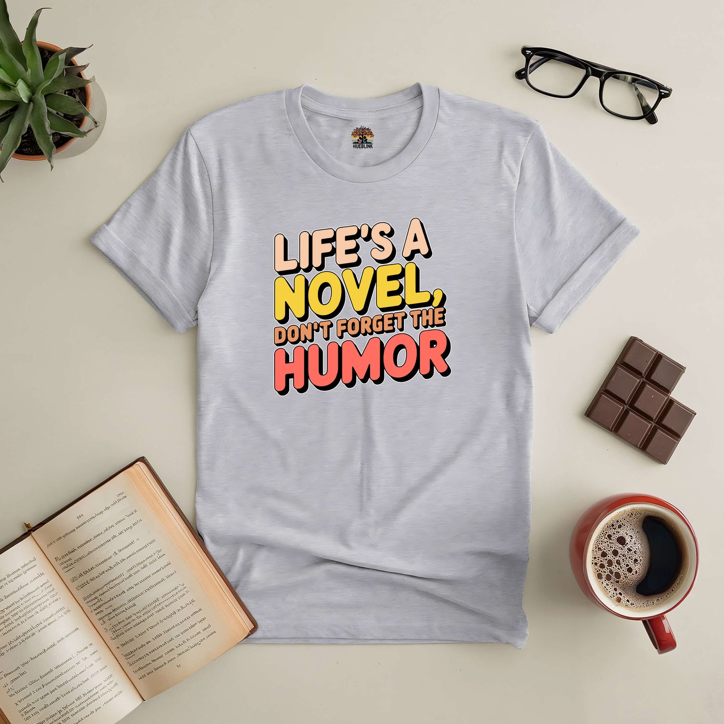 Gray tee with "Life's a Novel, Don't Forget the Humor" text, surrounded by an open book, glasses, chocolate, and coffee.