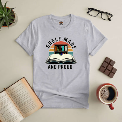 Shelf-Made And Proud tee for book lovers, featuring an open book design, surrounded by glasses, chocolate, coffee, and plants.