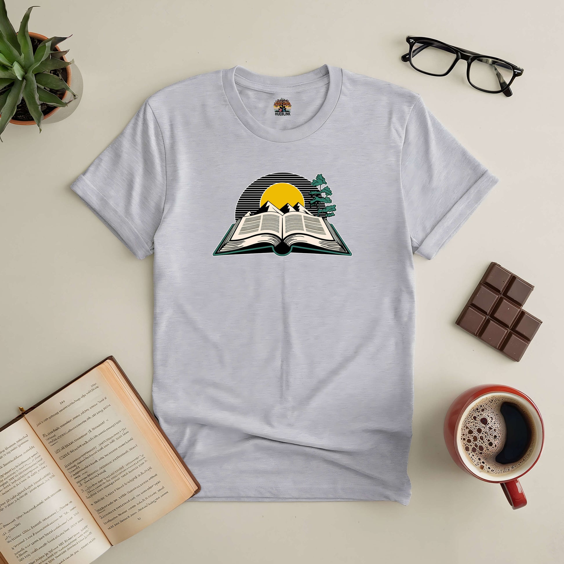 "Where Every Story Shines Tee with open book and sunrise graphic, surrounded by coffee and glasses, perfect for book lovers"