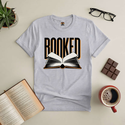 Booked tee featuring open book graphic, perfect for book lovers and busy minds, surrounded by coffee, glasses, and chocolate.