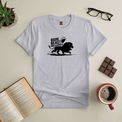 White "Book Beast Unleashed" tee with lion design, surrounded by book, coffee, glasses, and chocolate on a table.