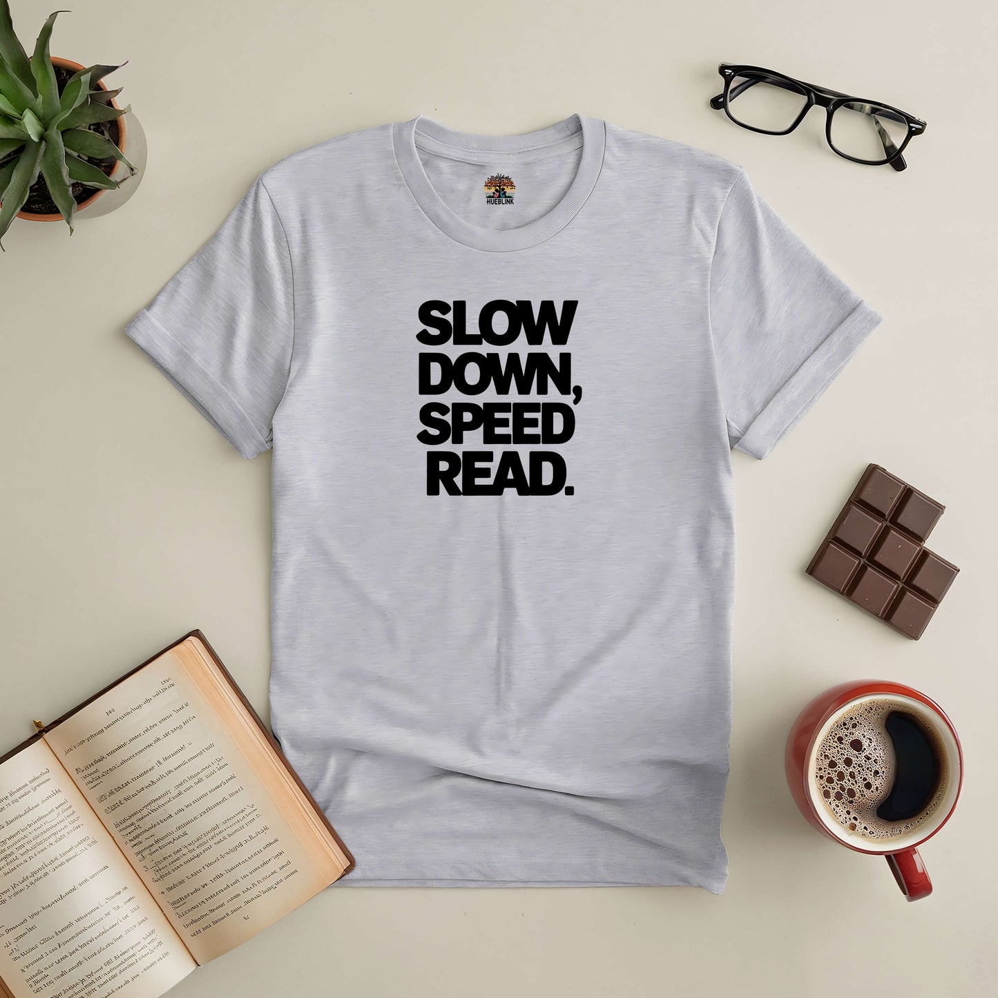 Grey tee with "Slow Down, Speed Read" design; surrounded by a book, coffee, glasses, chocolate, and plant on a beige background.