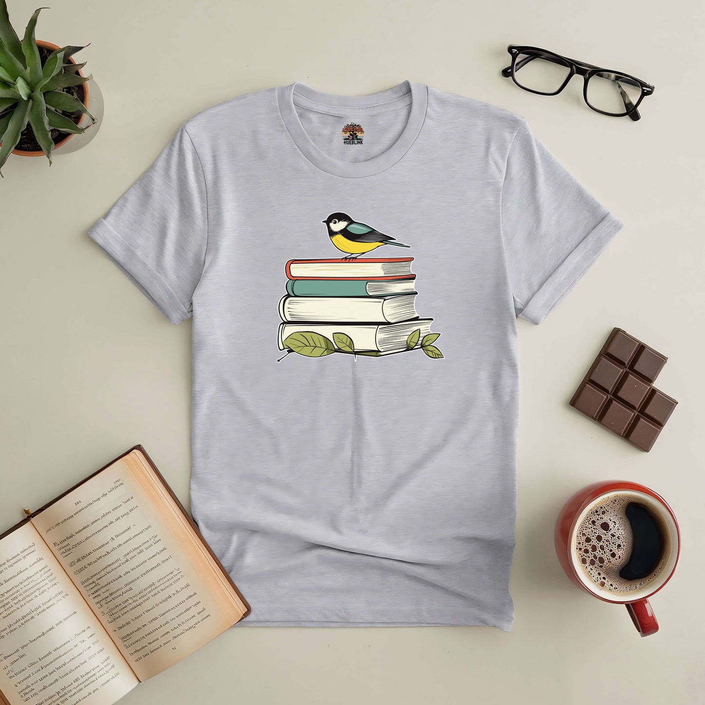 T-shirt featuring a bird on stacked books, surrounded by coffee, glasses, chocolate, and an open book. "Nestled in Stories" design.