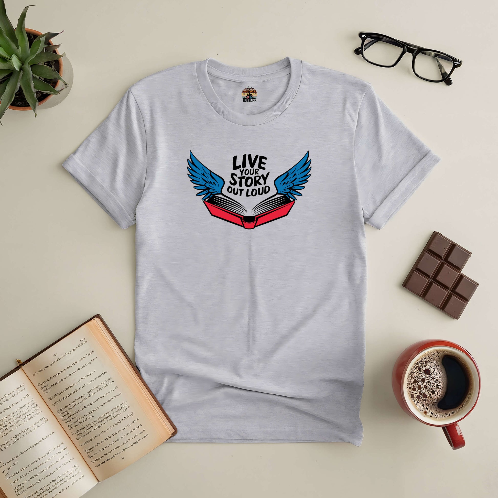 Live Your Story Out Loud Tee with open book and wings, perfect for dreamers and storytellers, surrounded by coffee and books.