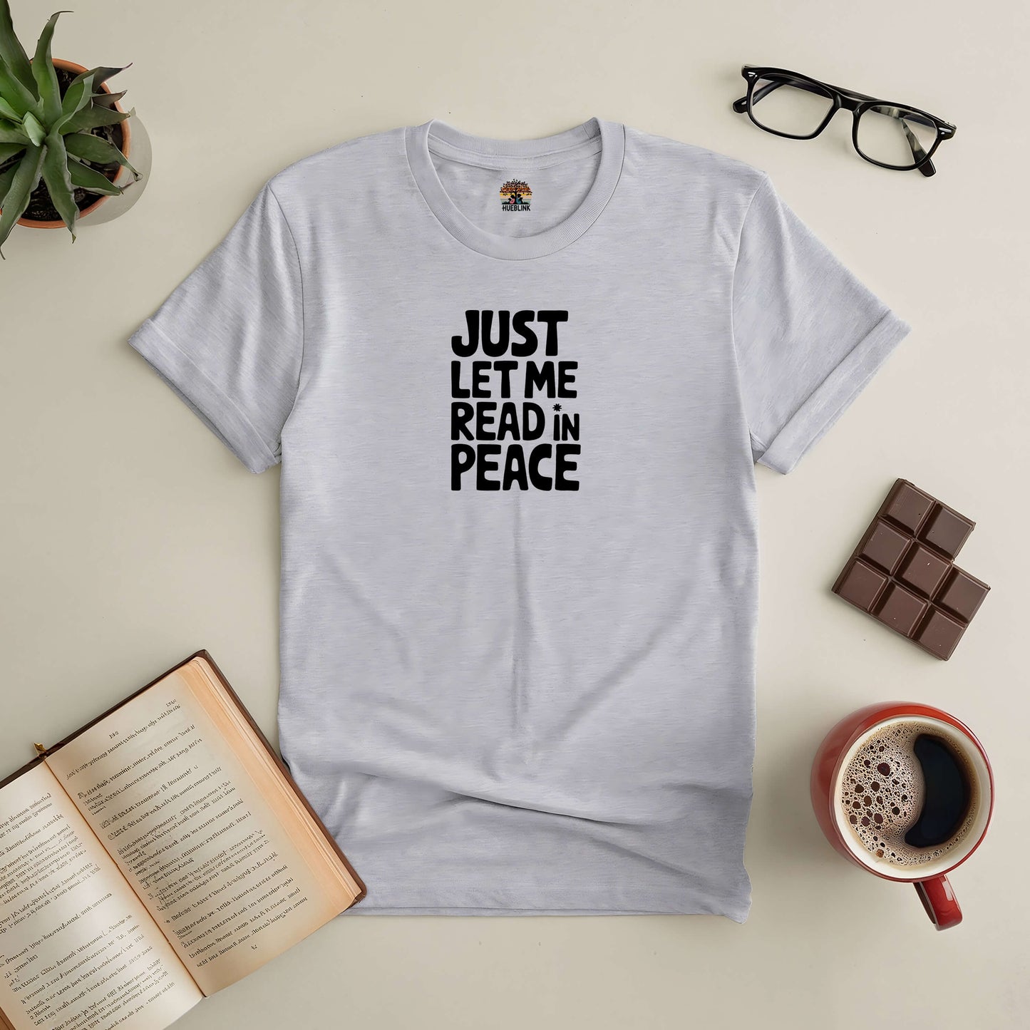 Grey t-shirt with "Just Let Me Read in Peace" text, laid on a table with an open book, glasses, chocolate, and a coffee mug.