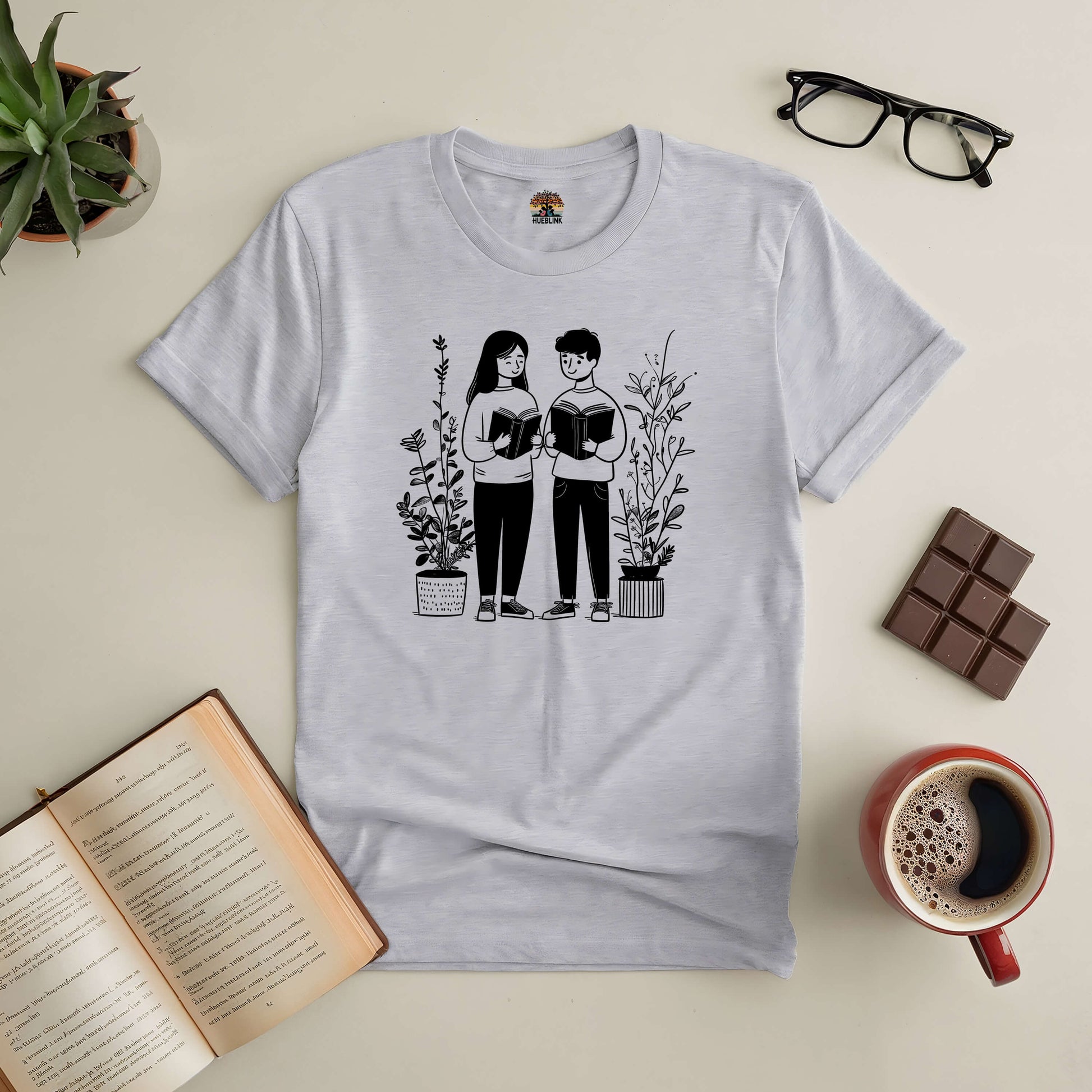 "Bookish Buddies Tee featuring two friends reading, perfect for literary duos, with books, plants, and coffee setup."