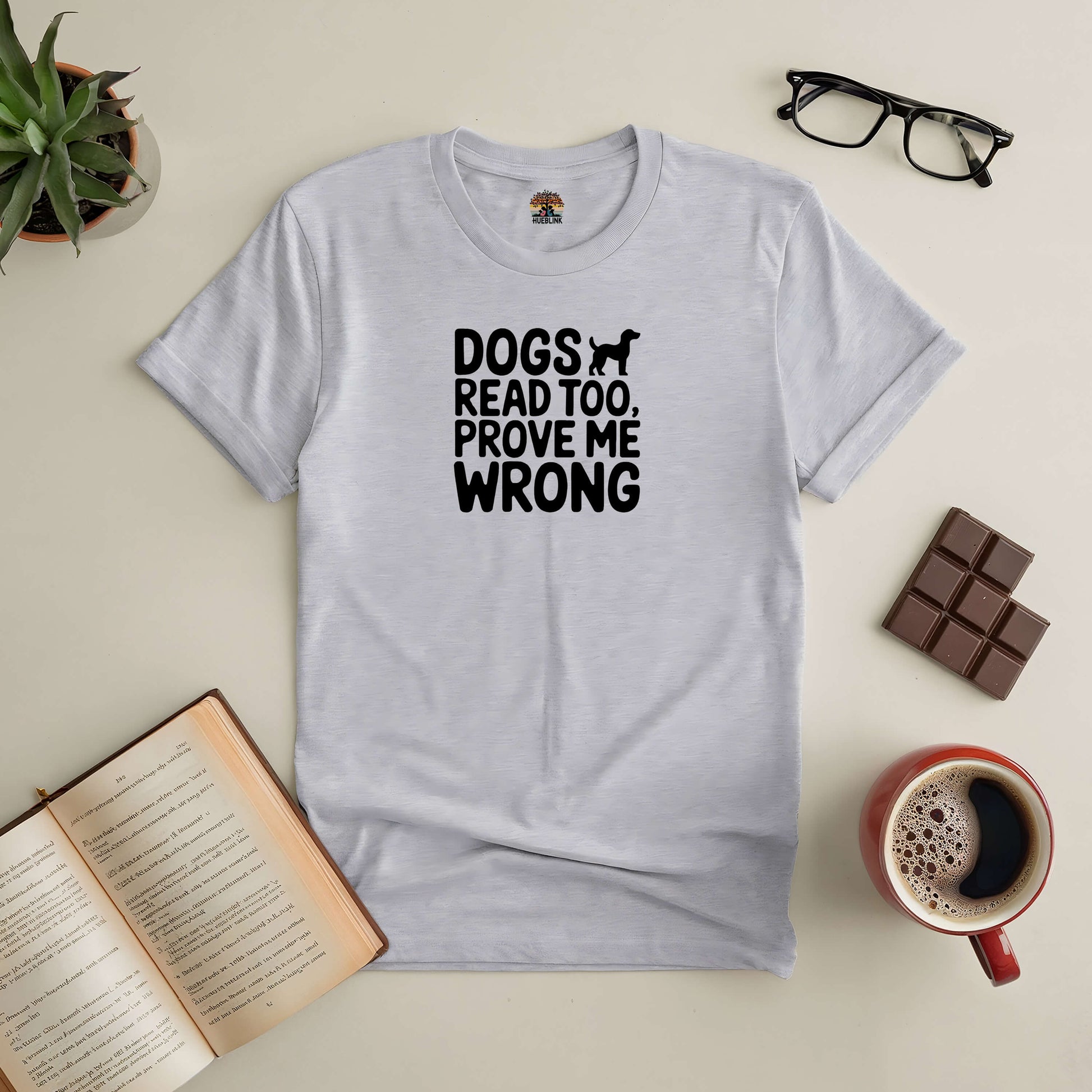 Gray tee with "Dogs Read Too, Prove Me Wrong" text, surrounded by an open book, coffee, chocolate, glasses, and succulent.