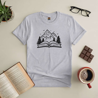 Gray "Peaks of Possibility" tee with mountain and book design, surrounded by coffee, chocolate, and glasses on a table.