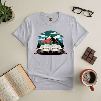 White tee with mountain and book graphic, surrounded by glasses, chocolate, a coffee cup, and an open book on a table.