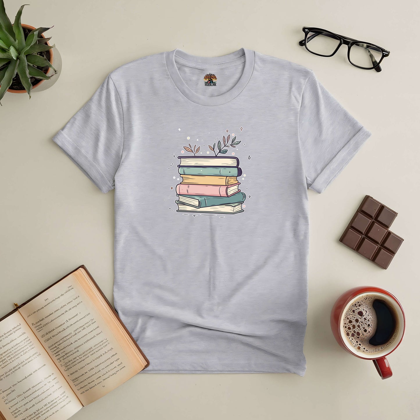 Blooming Books Tee with pastel-colored books and floral accents, surrounded by an open book, coffee, glasses, and chocolate.