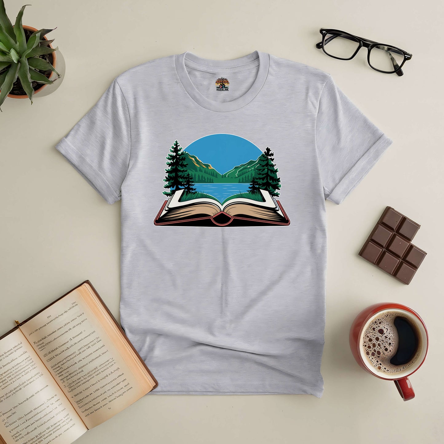 Book Your Escape tee with open book and nature scene, surrounded by coffee, chocolate, glasses, and an open book. Perfect for adventurers.