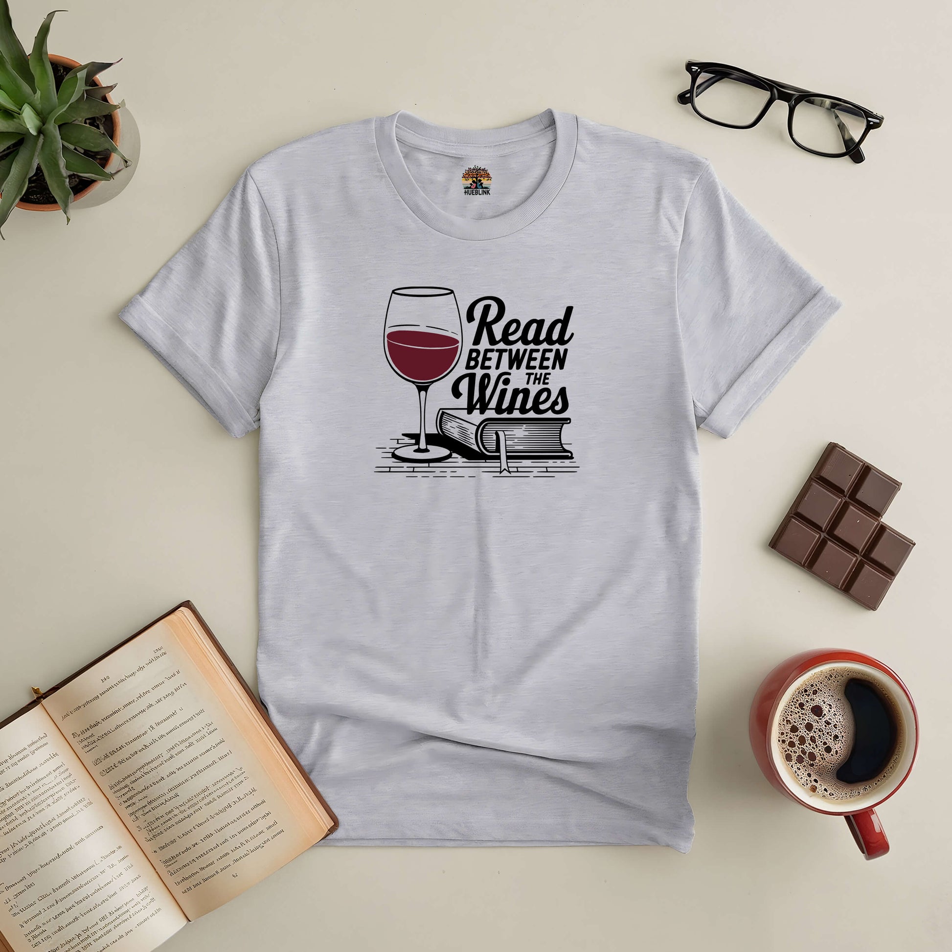 "Read Between the Wines Tee with book, wine glass, open book, and coffee on a table for book and wine lovers"