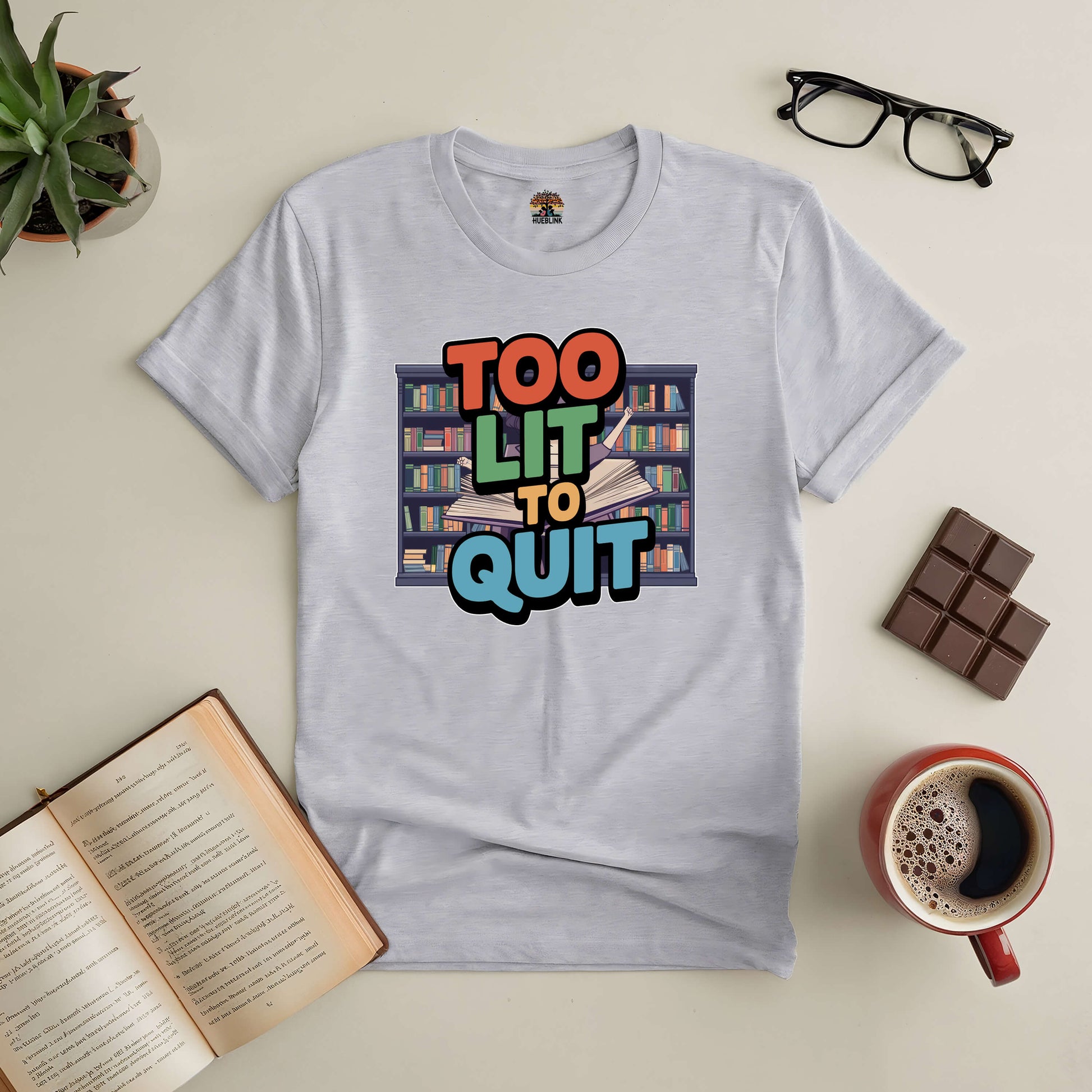 White tee with "Too Lit To Quit" design, surrounded by books, chocolate, coffee, and reading glasses on a cozy setup.