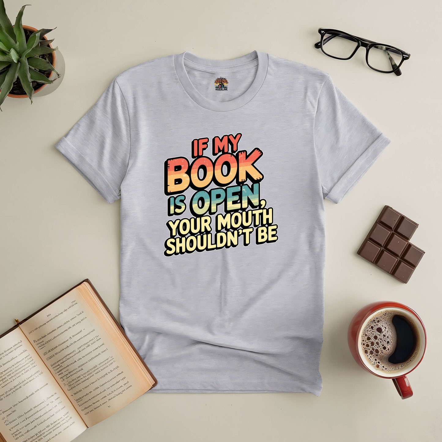 Gray tee with text "If My Book Is Open, Your Mouth Shouldn't Be" surrounded by an open book, glasses, chocolate, and coffee.