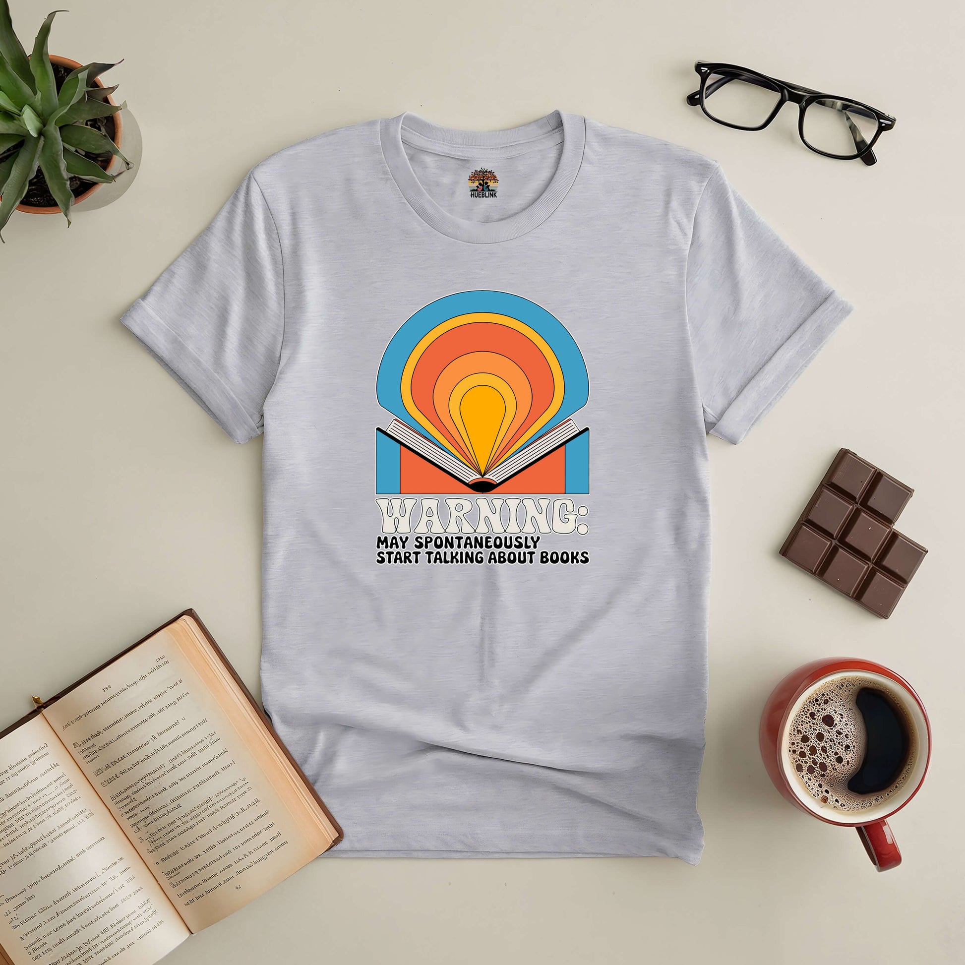 "Gray book-themed tee with colorful design, surrounded by open book, coffee, glasses, and chocolate, perfect for book lovers."