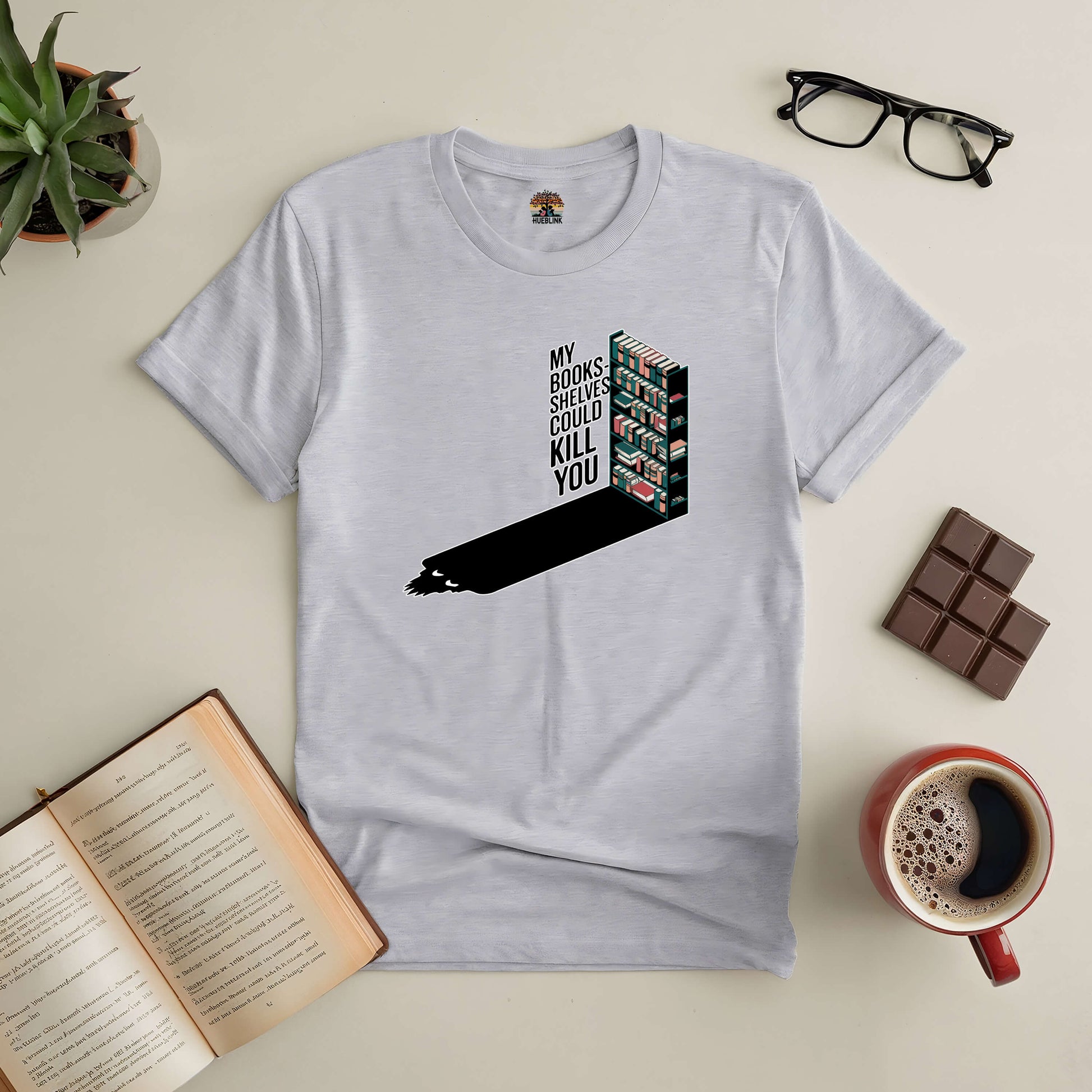 "Book-themed tee 'My Bookshelves Could Kill You' laid out with coffee, glasses, chocolate, and open book for book lovers"