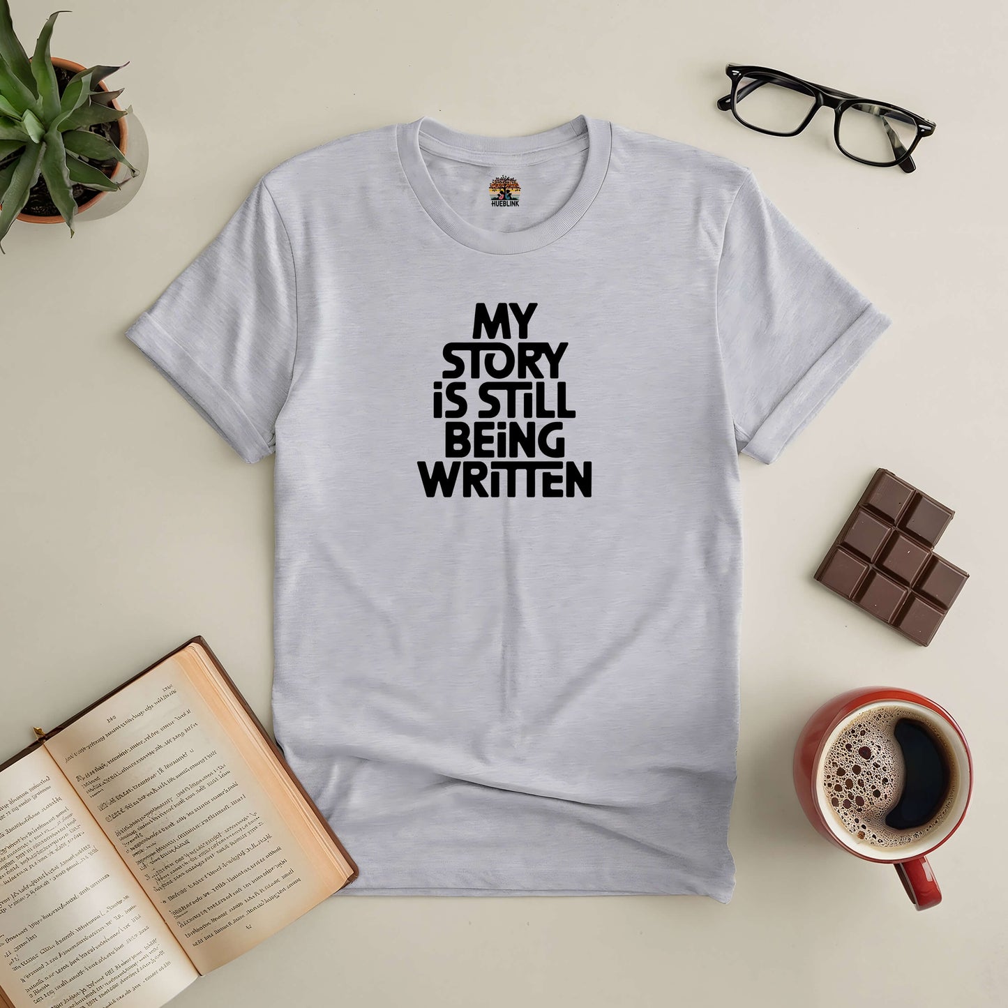 White t-shirt with "My Story Is Still Being Written" text, placed alongside a book, coffee, glasses, chocolate, and a plant.