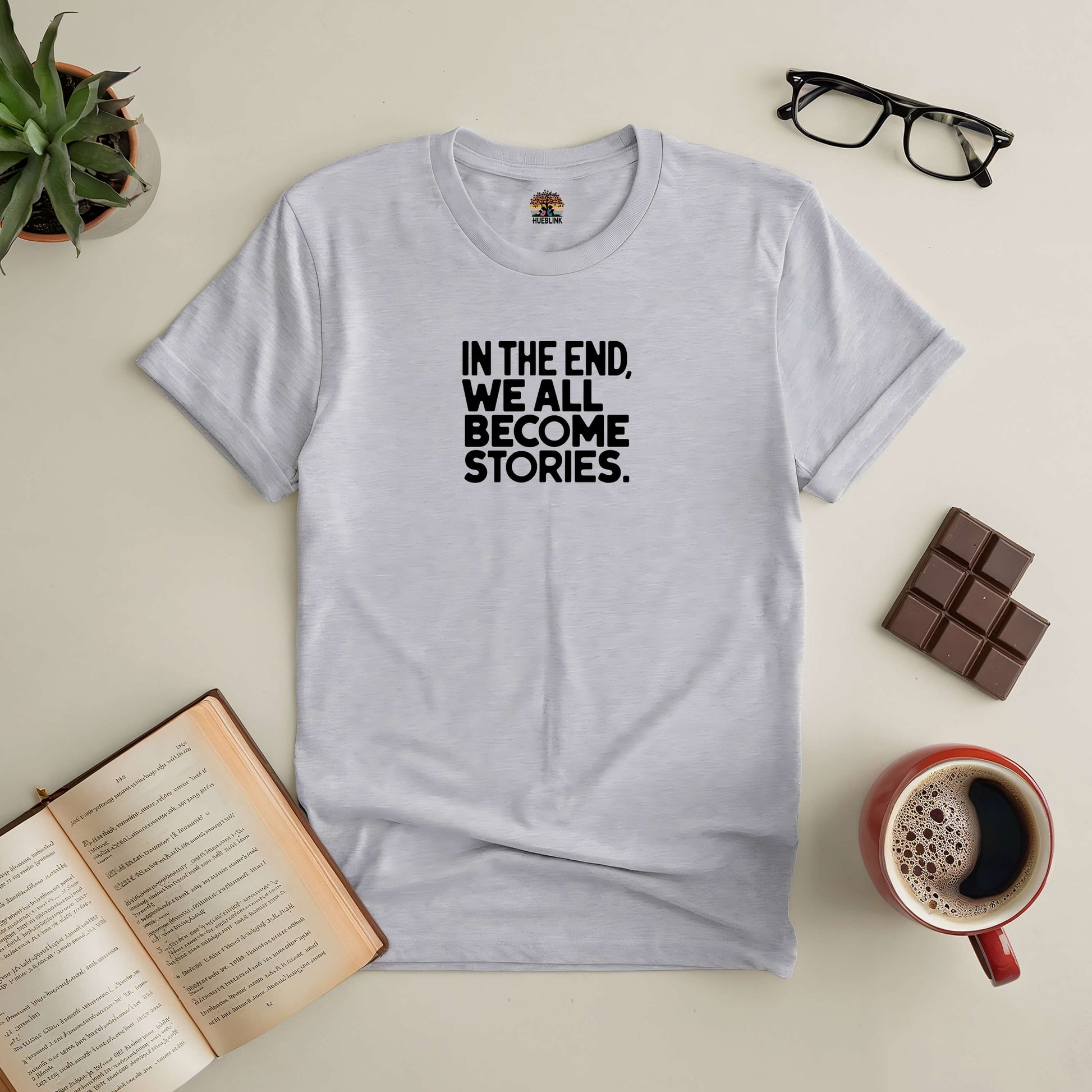 "In The End, We All Become Stories Tee laid out with book, coffee, and glasses on a table"