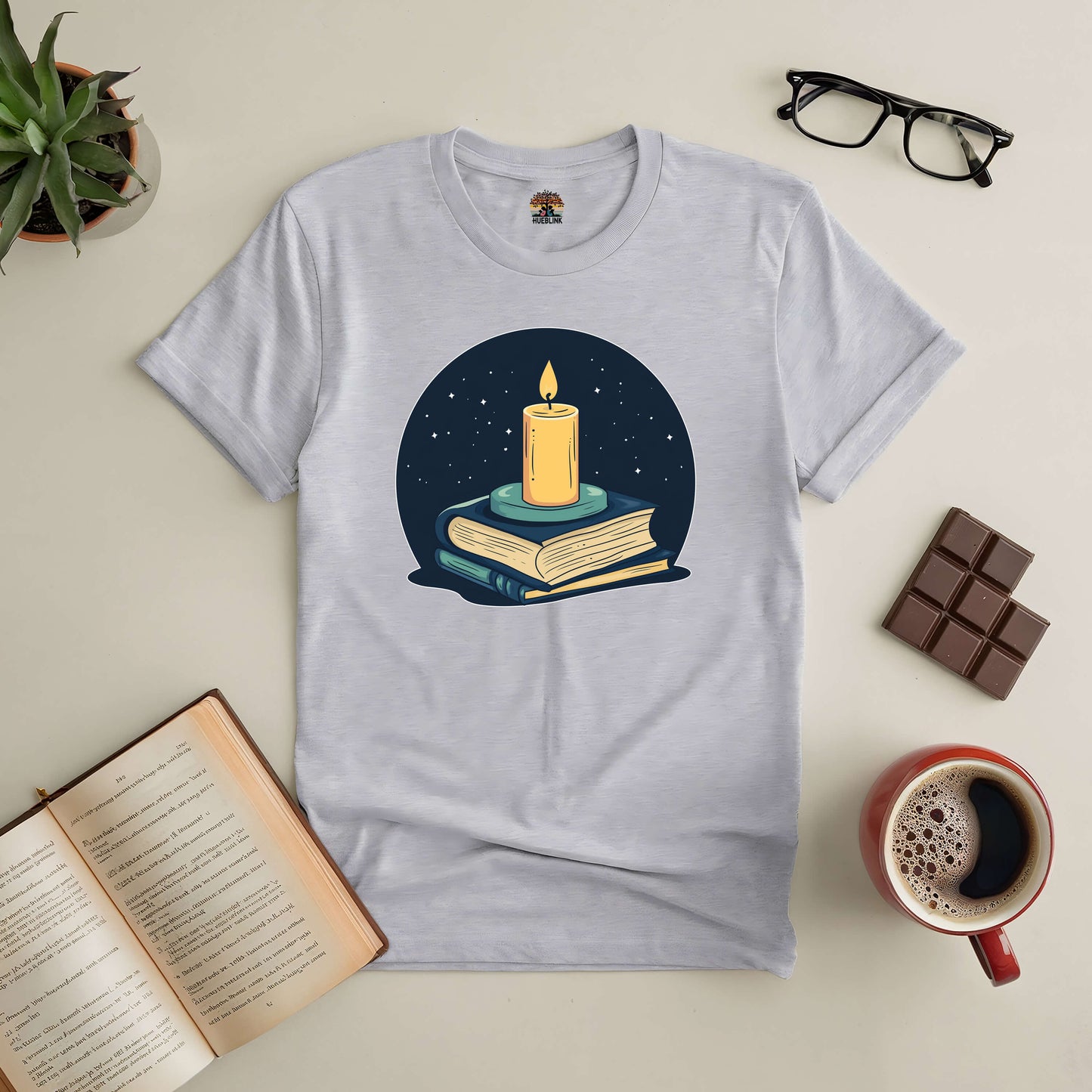 "Illuminate Your Mind Tee with candle and books design, surrounded by coffee, chocolate, and open book on a table setting."