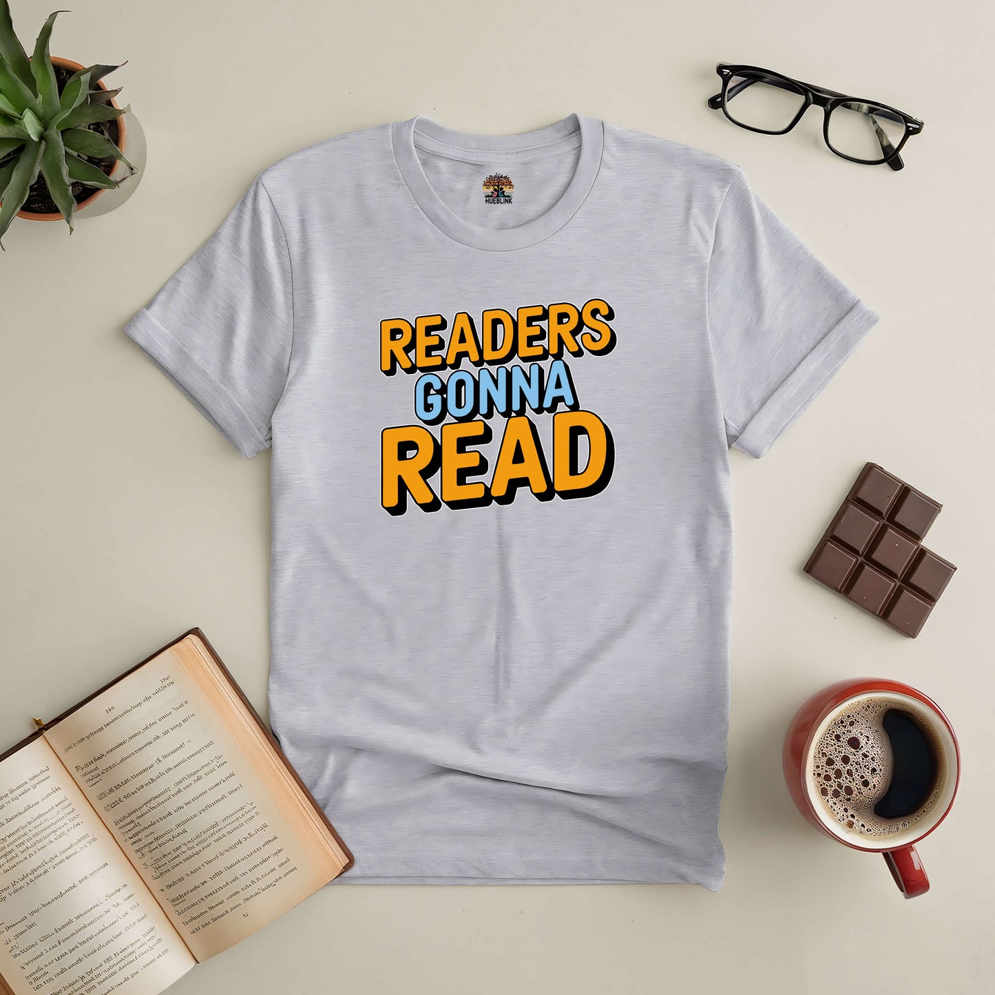 White "Readers Gonna Read" tee with colorful text, placed on a table with an open book, glasses, chocolate, and a coffee cup.