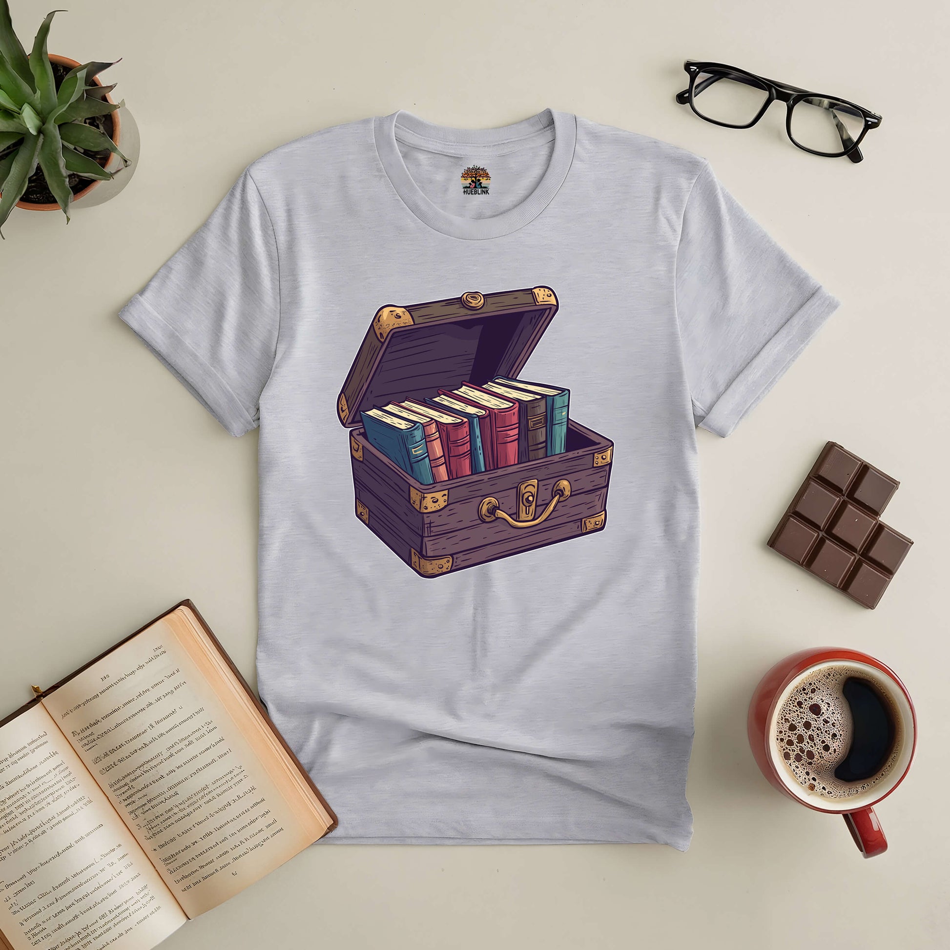 "Priceless Treasure Tee with book-filled chest design for book lovers, surrounded by an open book, glasses, plant, chocolate, and coffee."