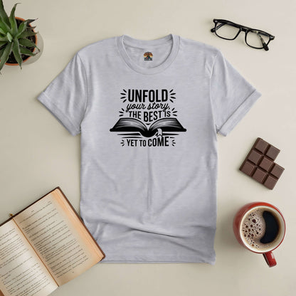 Motivational tee with "Unfold Your Story" design, featuring an open book. Displayed on a flat surface with coffee, glasses, and chocolate.