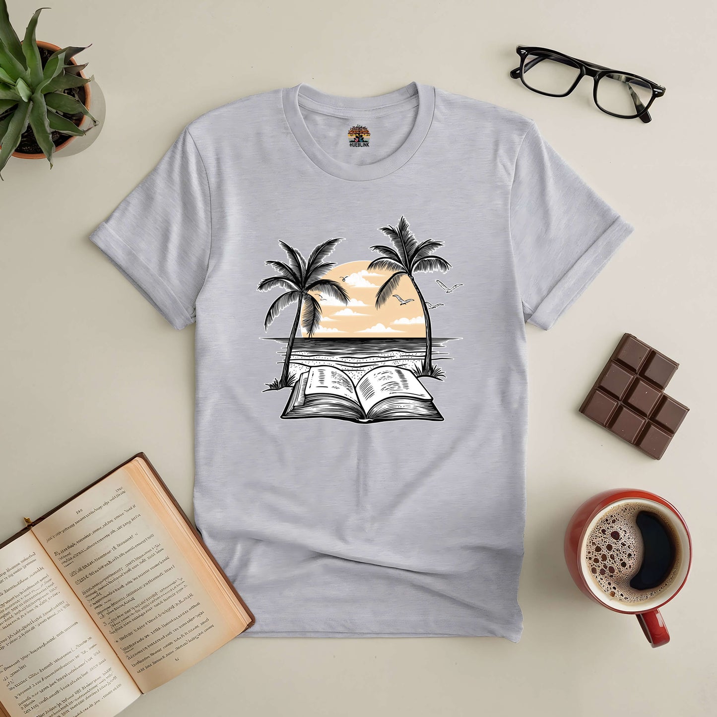 Beachside Bibliophile Tee with open book and palm tree design, surrounded by coffee, chocolate, glasses, and a plant.