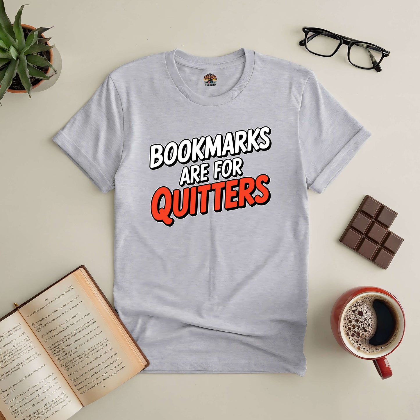 "Bookmarks Are for Quitters tee for avid readers, featuring bold lettering on a gray t-shirt, surrounded by a book, coffee, and chocolate."