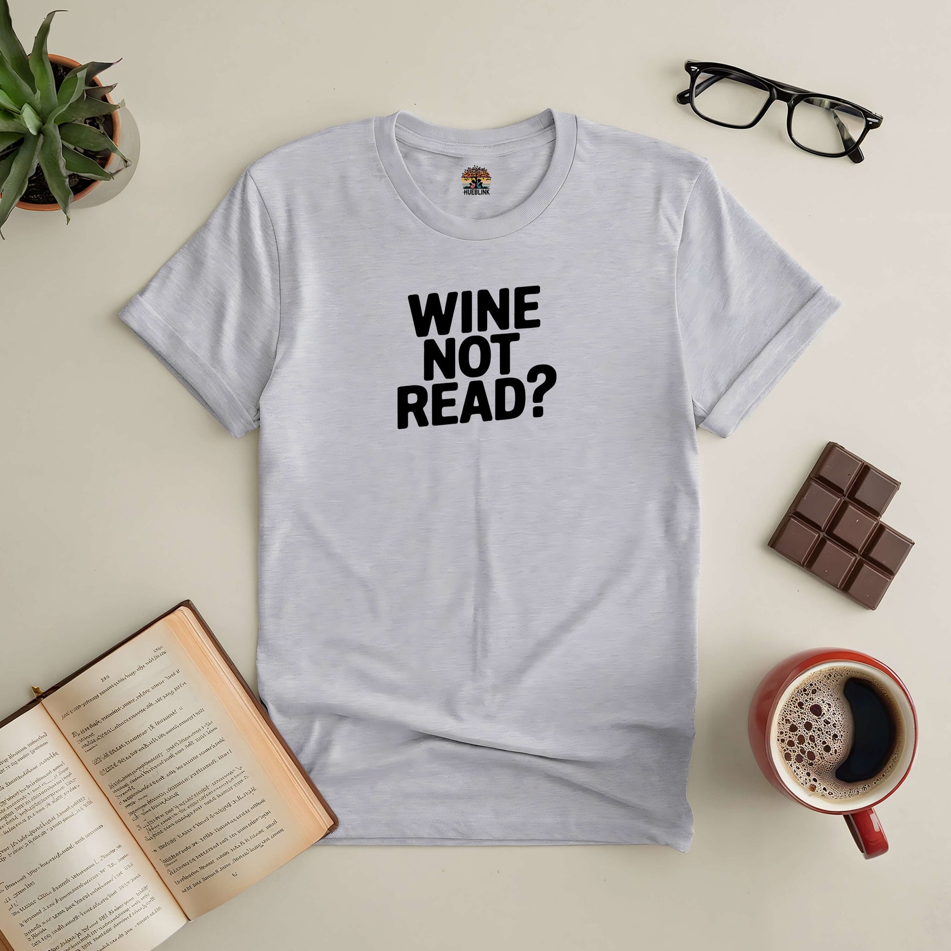 Gray tee with "Wine Not Read?" text, surrounded by book, glasses, chocolate, coffee, and plant for wine-loving bookworms.