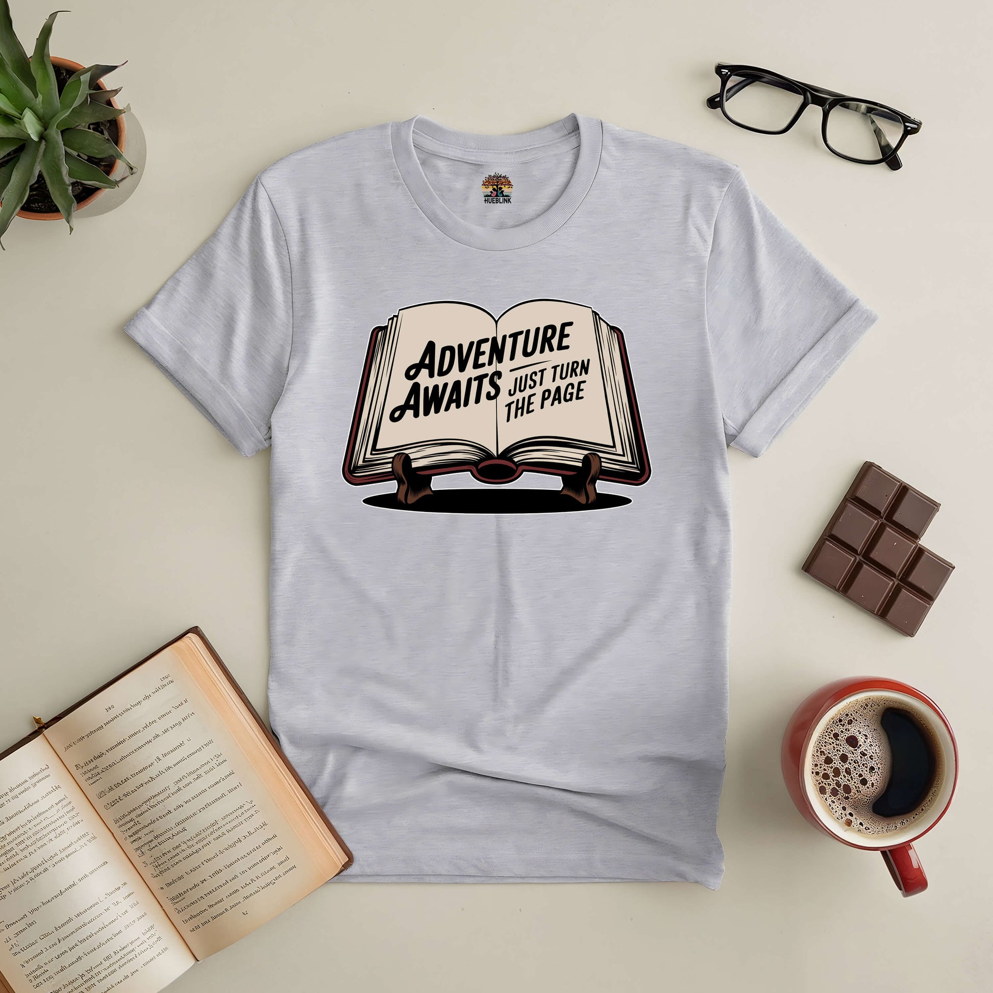 "Adventure Awaits tee with book design, surrounded by coffee, chocolate, glasses, and plants, inspiring new journeys."